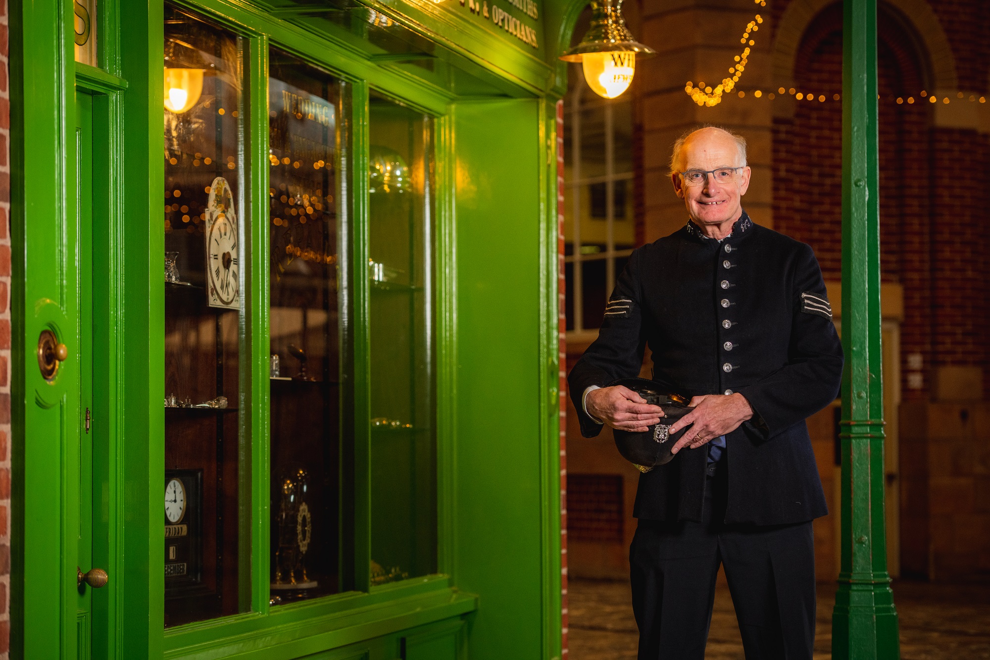 A man smiles at the camera, he has short hair and wears thin black rimmed glasses. He is dressed in a WWII policeman outfit and stands infront of a green shop front.