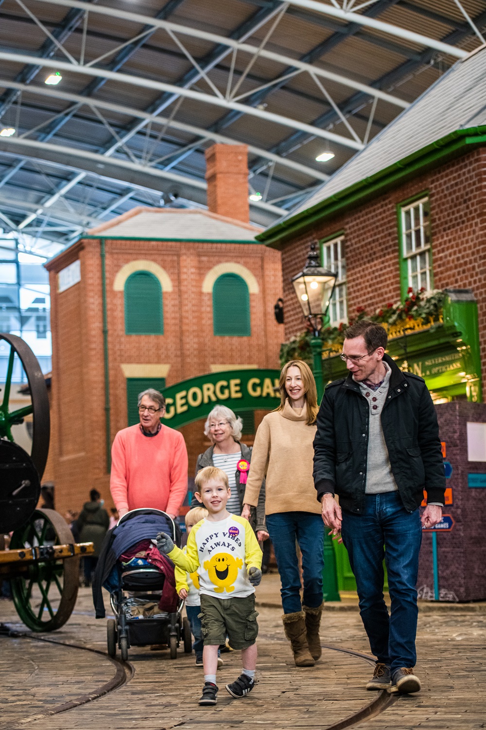 A group walks towards the camera, they walk on cobbled stones. There are six people within the image. To the front of the group is a small boy wearing a white top with a large yellow smiley face.  A man walks next to him wearing a black jacket and blue jeans. Behind them in the group is a lady wearing a brown jumper and blue jeans. A man pushes a pram wearing a salmon coloured jumper. Next to him is a lady with white hair wearing a striped top. Slightly cropped to the left of the image is a victorian machine.