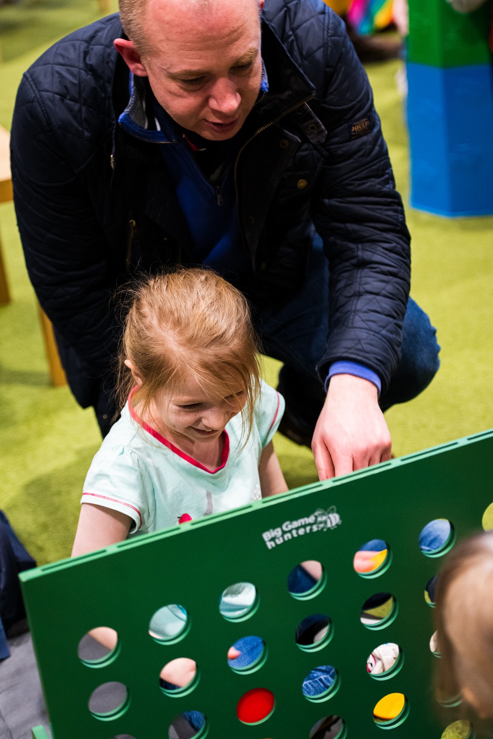 A young girl in a green top plays a large Connect 4 game using red and yellow discs. A man kneels next to her wearing a navy blue jacket and blue jeans.