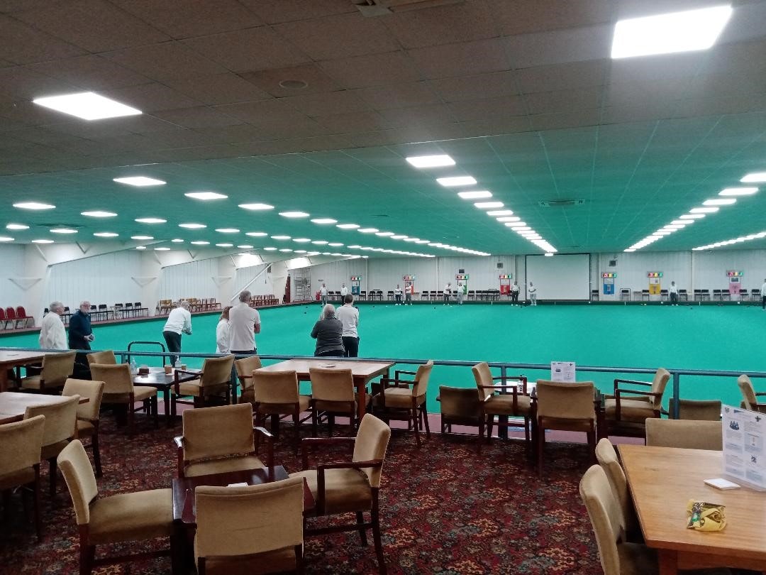 An indoor bowling green with a collection of people playing. In front of the pitch you can see tables and chairs.