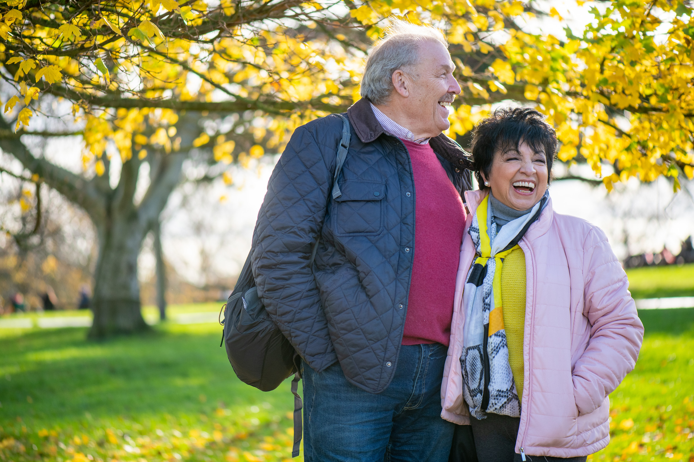 An older gentleman stands next to an older female they are in the park.