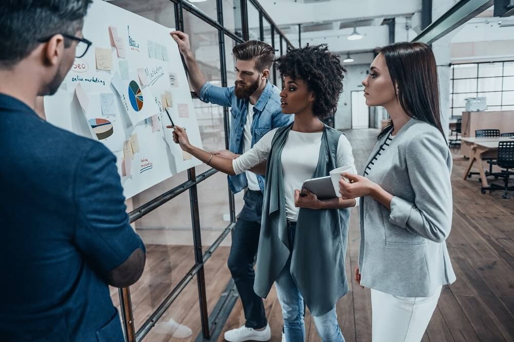 A group of 4 people stand looking at a board which has post it notes and graphs on.