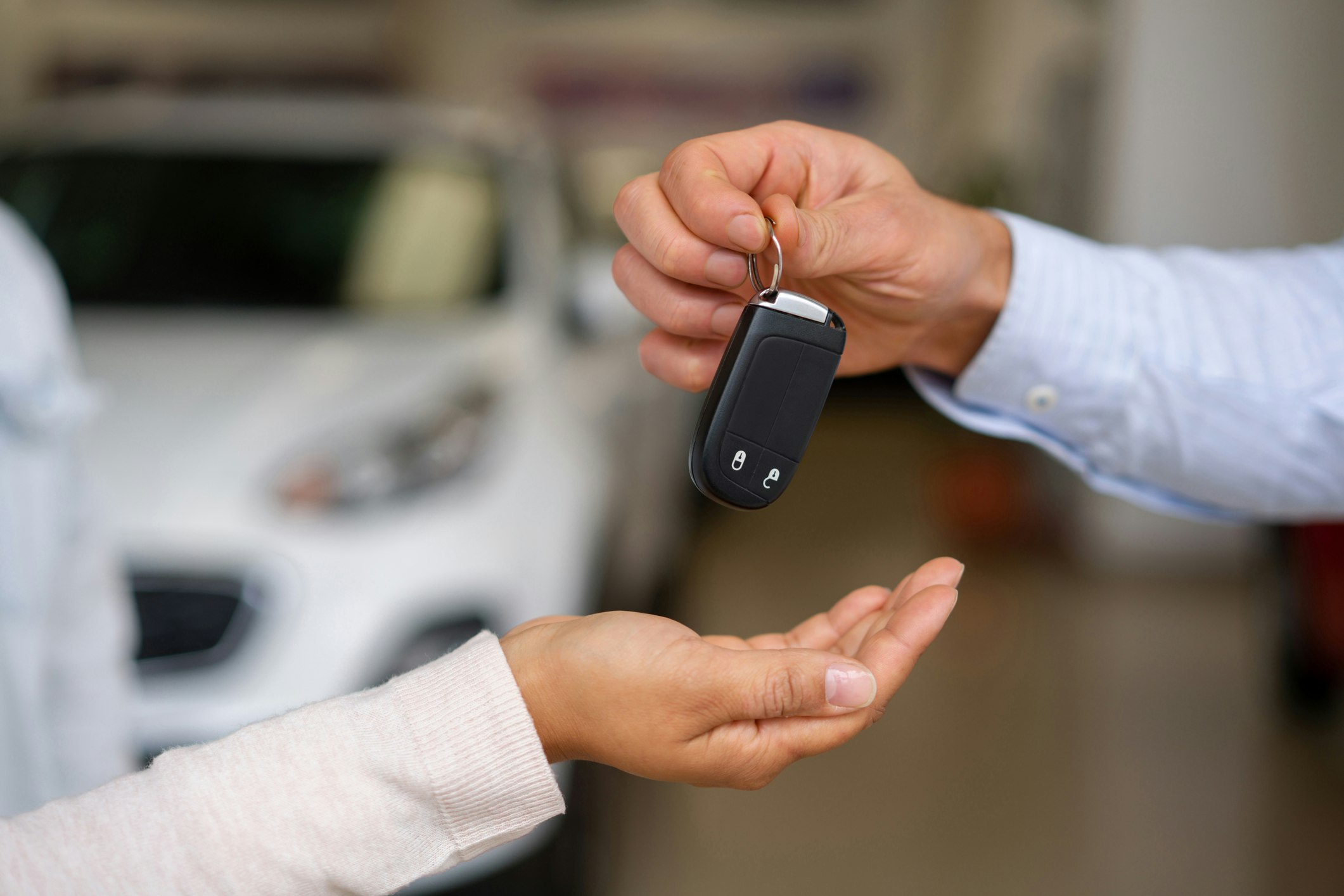 A person hands over a car key to another who has their hand cupped and under the key. In the background you can see a blurry image of a car.