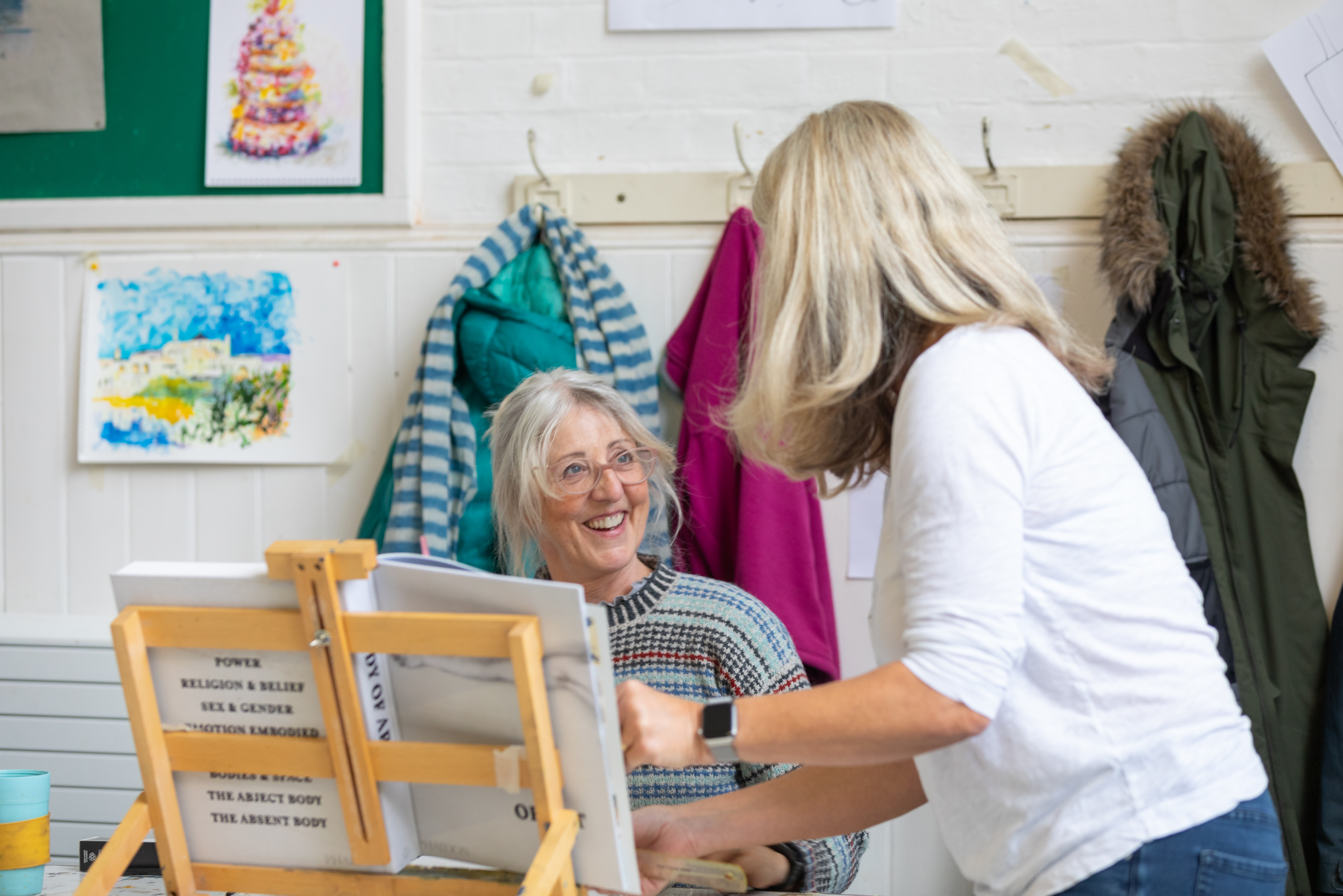 A lady is smiling and sitting behind a wooden easel that has a book on it. Another lady is talking to her.