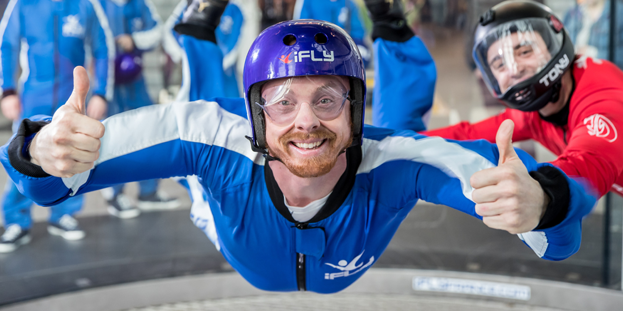 A man wearing a jump suit and a helmet is floating in mid air with his arms to the side and thumbs up. He looks at the camera. To his side is an instructor wearing a red jumpsuit and black helmet.