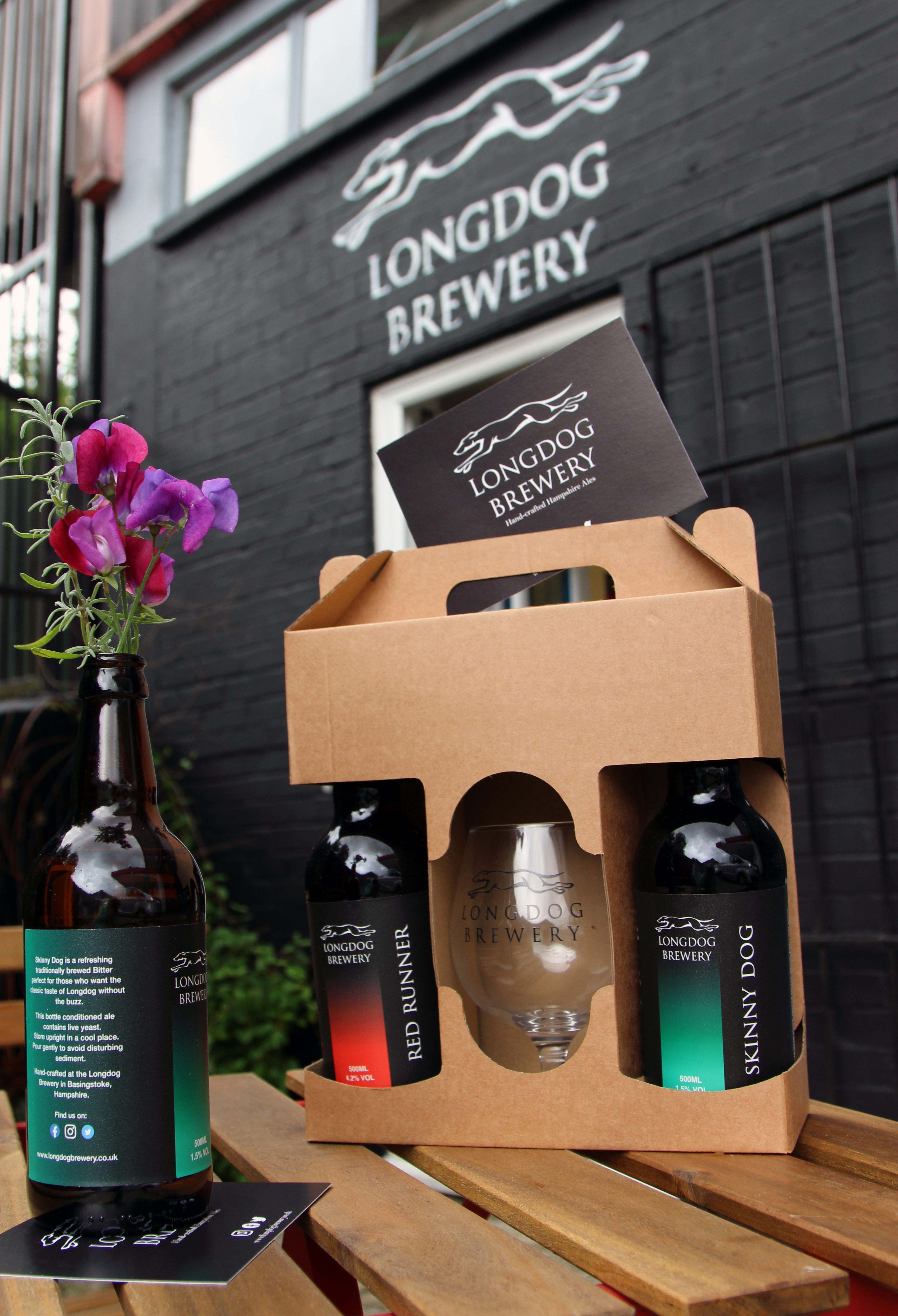 A box which has two bottles of beer and a glass. It's sat on a wooden table and has an open bottle of beer with flowers in.