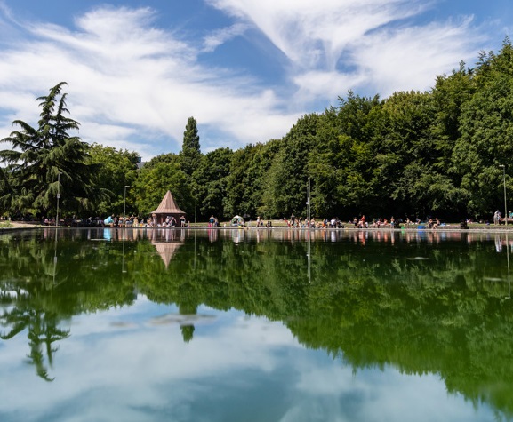 A landscape image showing a blue sky with white fluffy clouds and green trees are reflected on a pond. Horizontal across the middle of the image are groups of people relaxing and having fun in the park.