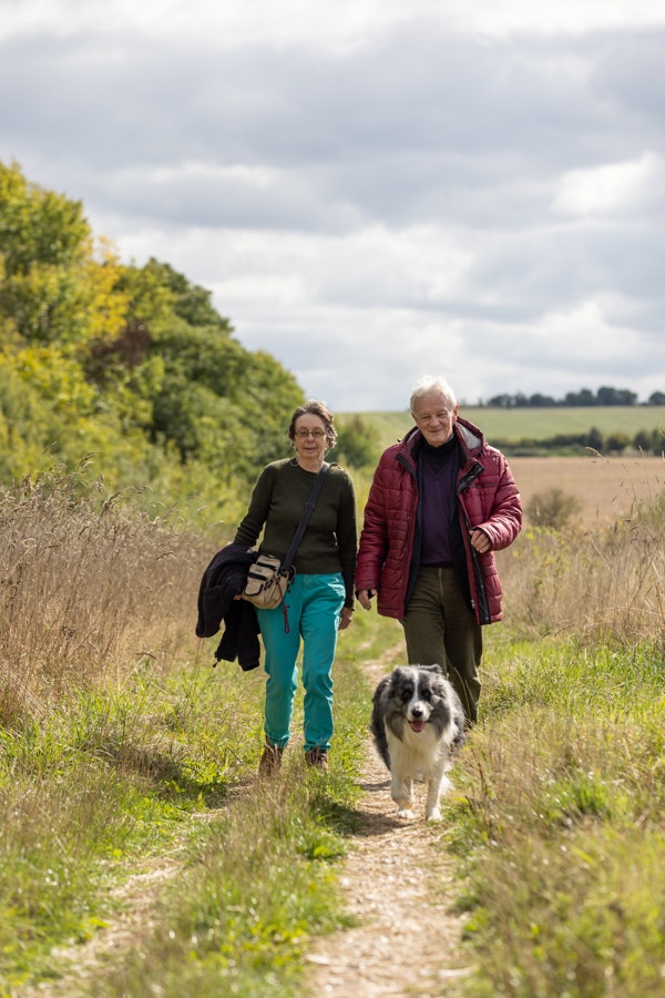 Two people walk with their dog in the Countryside. A natural path is created through the grass. The lady wears green trousers and a brown t-shirt, she carries her coat. The man wears a dark green trousers and a magenta puffa coat. A grey and white dog races towards the camera.