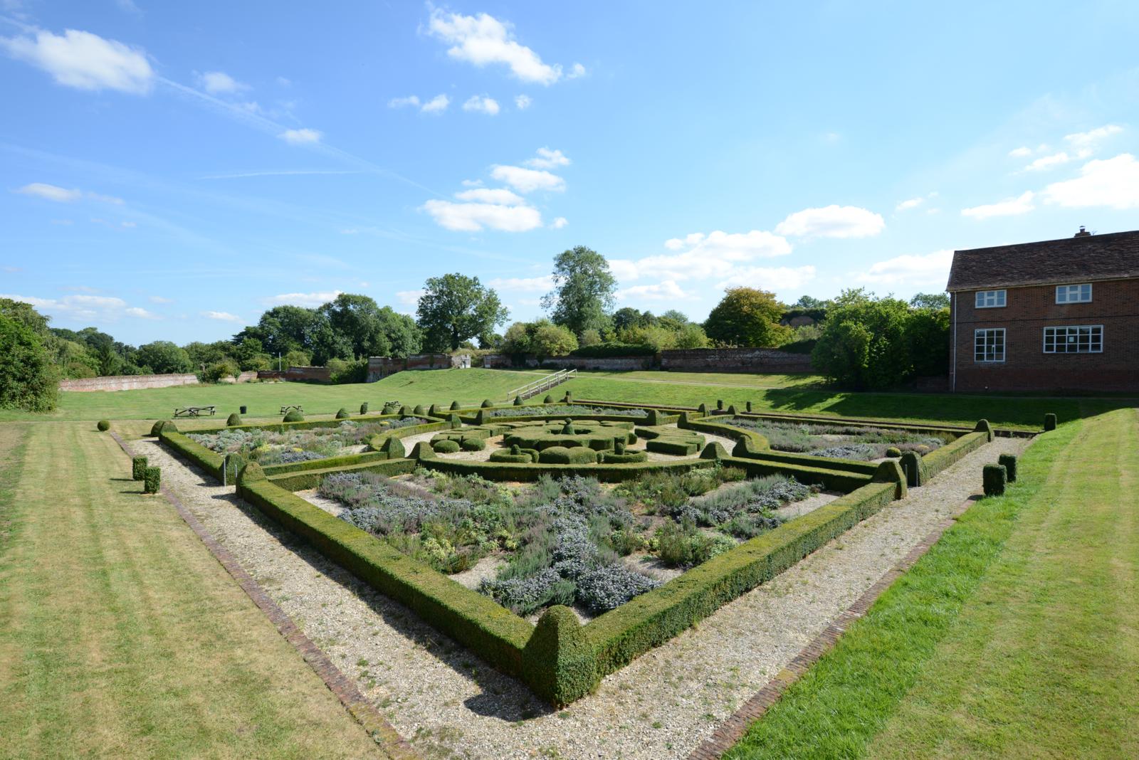 A green space with walls and a building. In the middle you can see a planted garden carefully curated with shrubs and flowers.