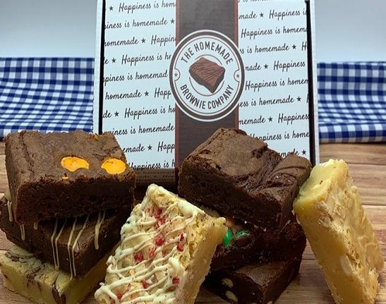 A selection of rectangular shaped brownies in front of a homemade brownie company gift box and a blue gingham table cloth in the background.