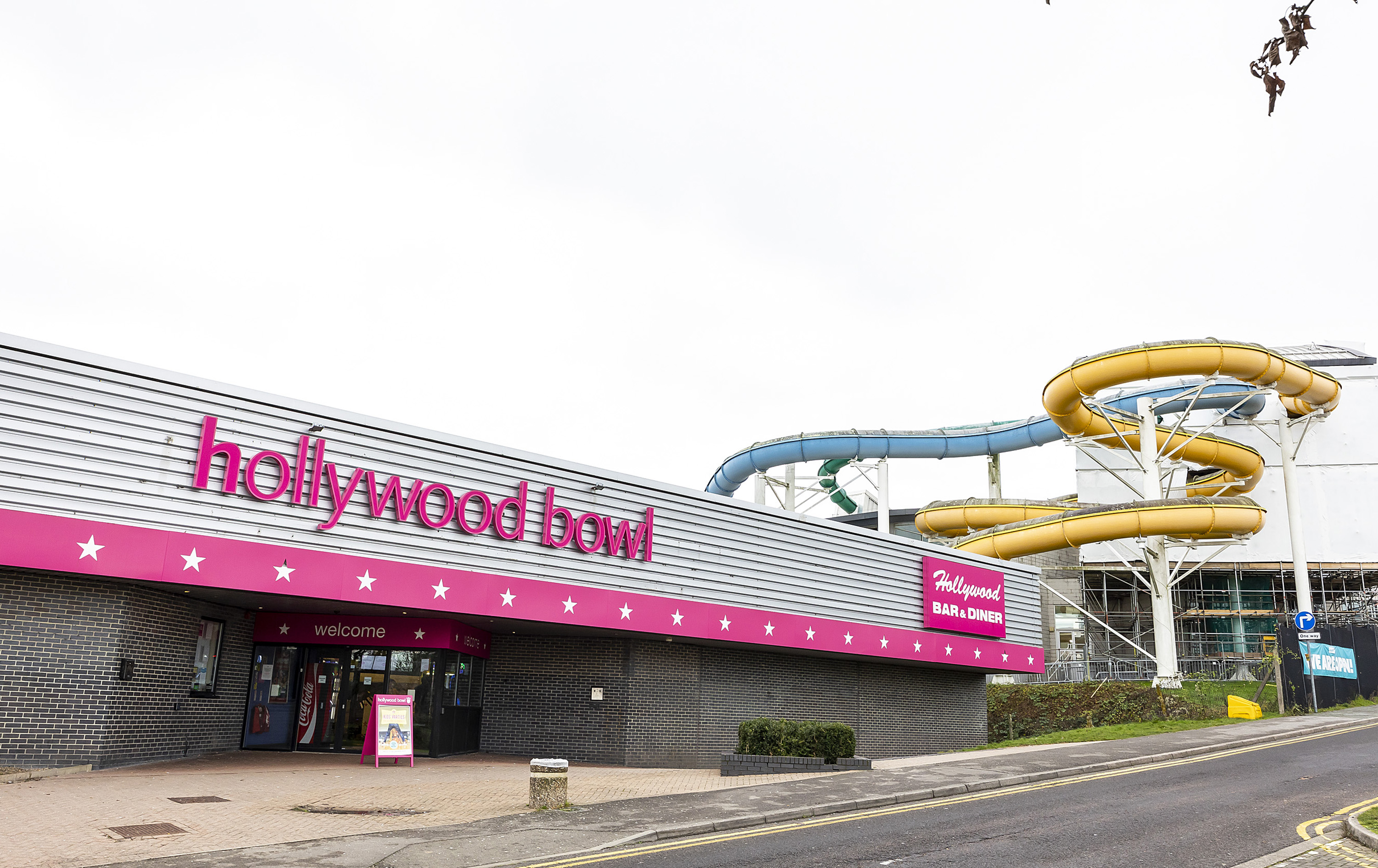 A brown brick building with a silver and pink facade. Hollywood bowl sign and a Bar and Diner sign. The pink park of the sign has white stars on. In the background you can see the flumes of the Aquadrome.