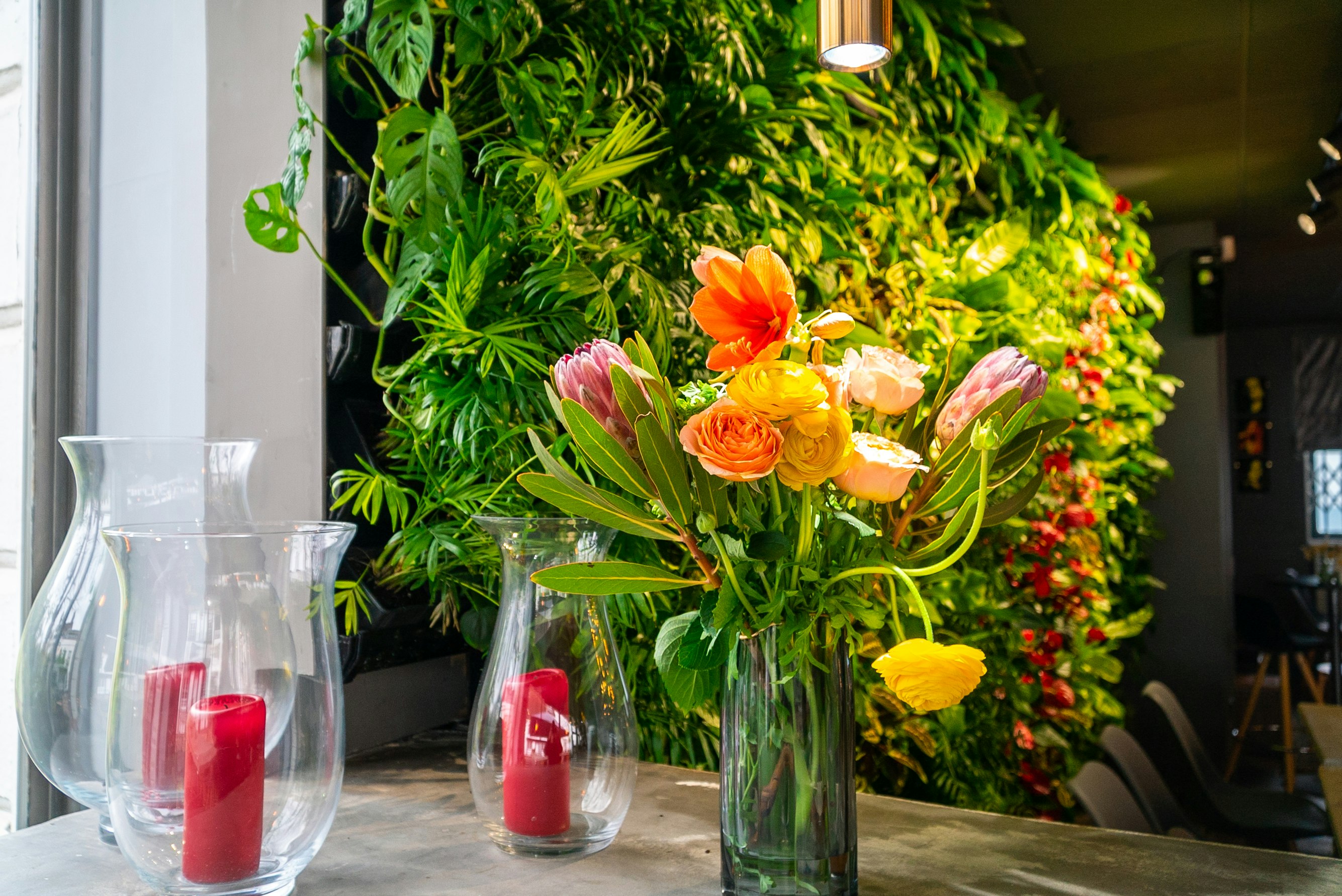 A table is pictured with a vase of flowers and three clear vases contain red candles. In the backdrop is a green living foliage wall inside a restaurant.