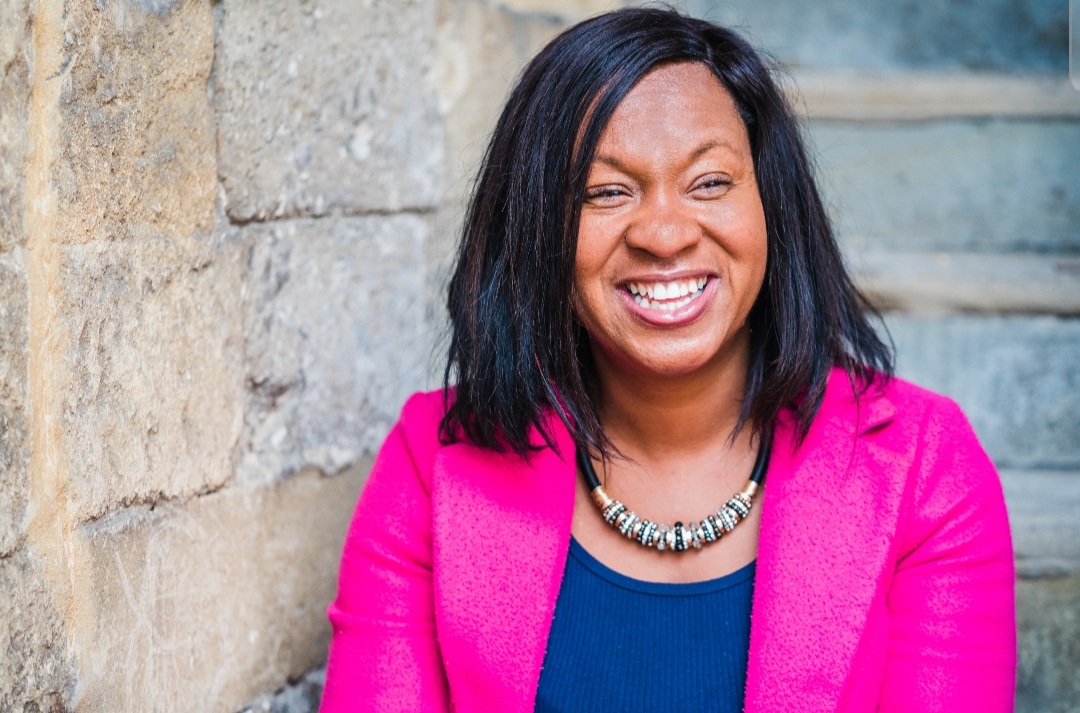 A women wearing a bright pink jacket is smiling and looking to the right. She stands against a brick wall.