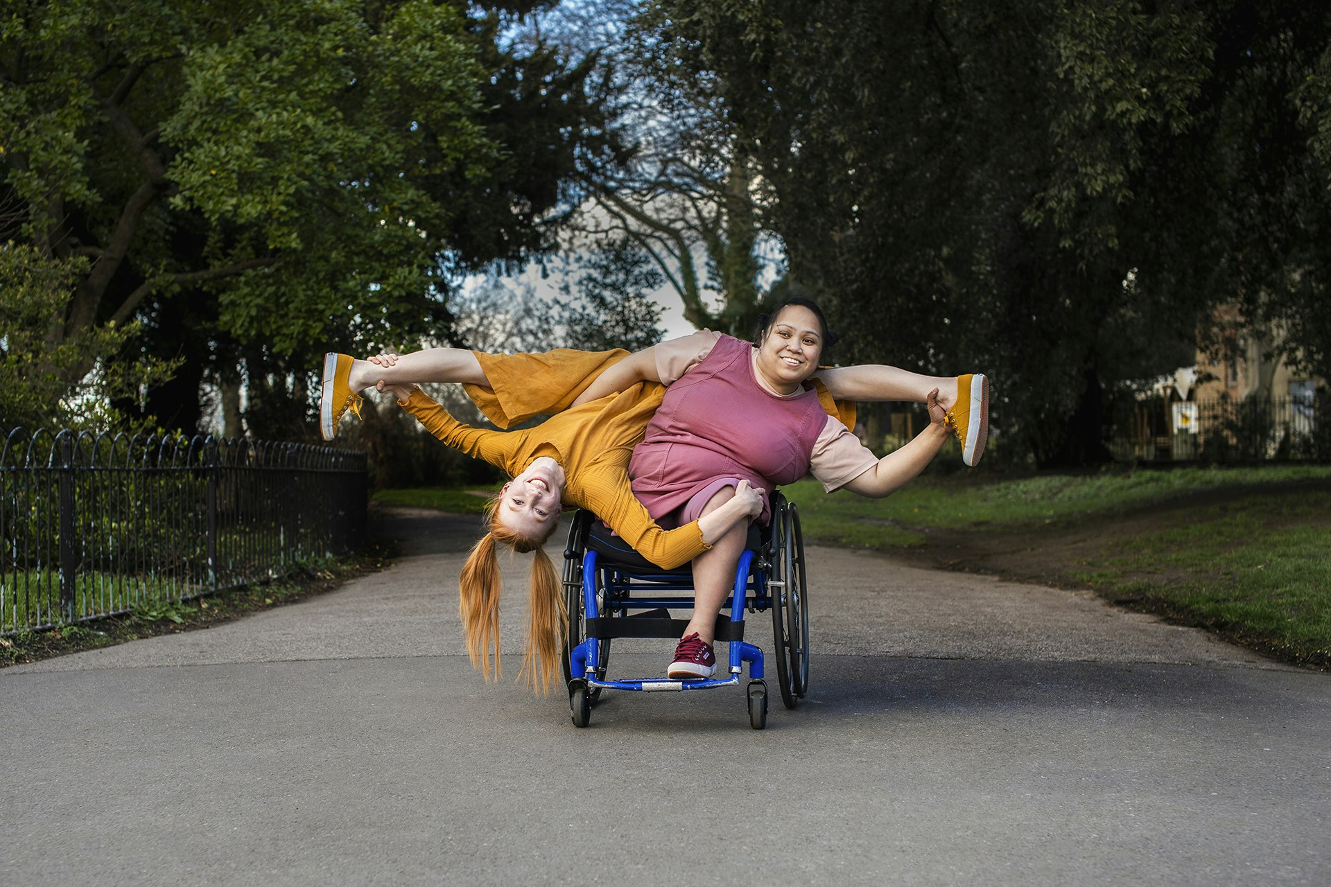 Two people smile at the camera, central to the image on a grey path. One person is a wheelchair dancer who sits in a blue wheelchair wearing purple. The other person wears a yellow dungarees and is upside down in the image. She works with the other performer to balance upside down on the wheelchair facing the camera.