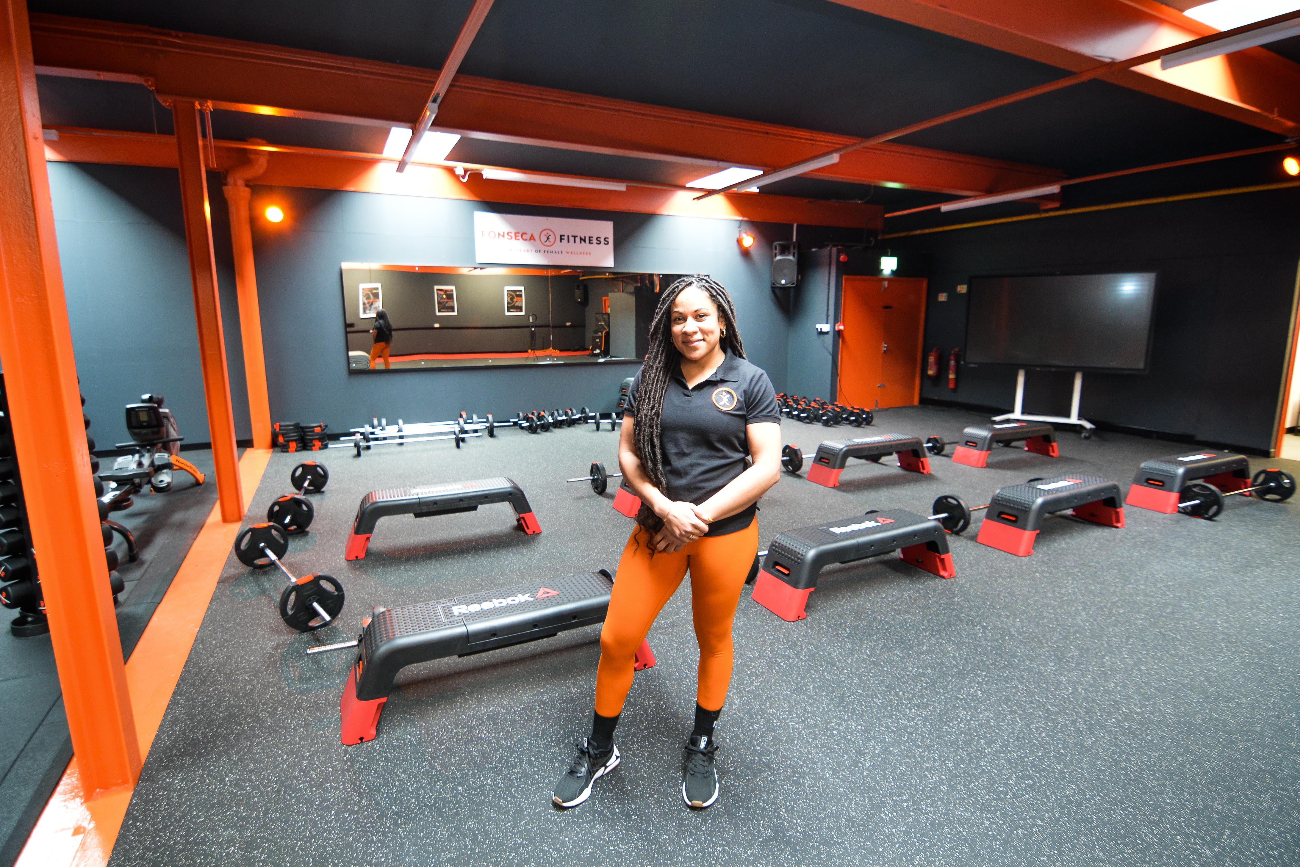 A lady stands in the gym. You can see weights and steps.