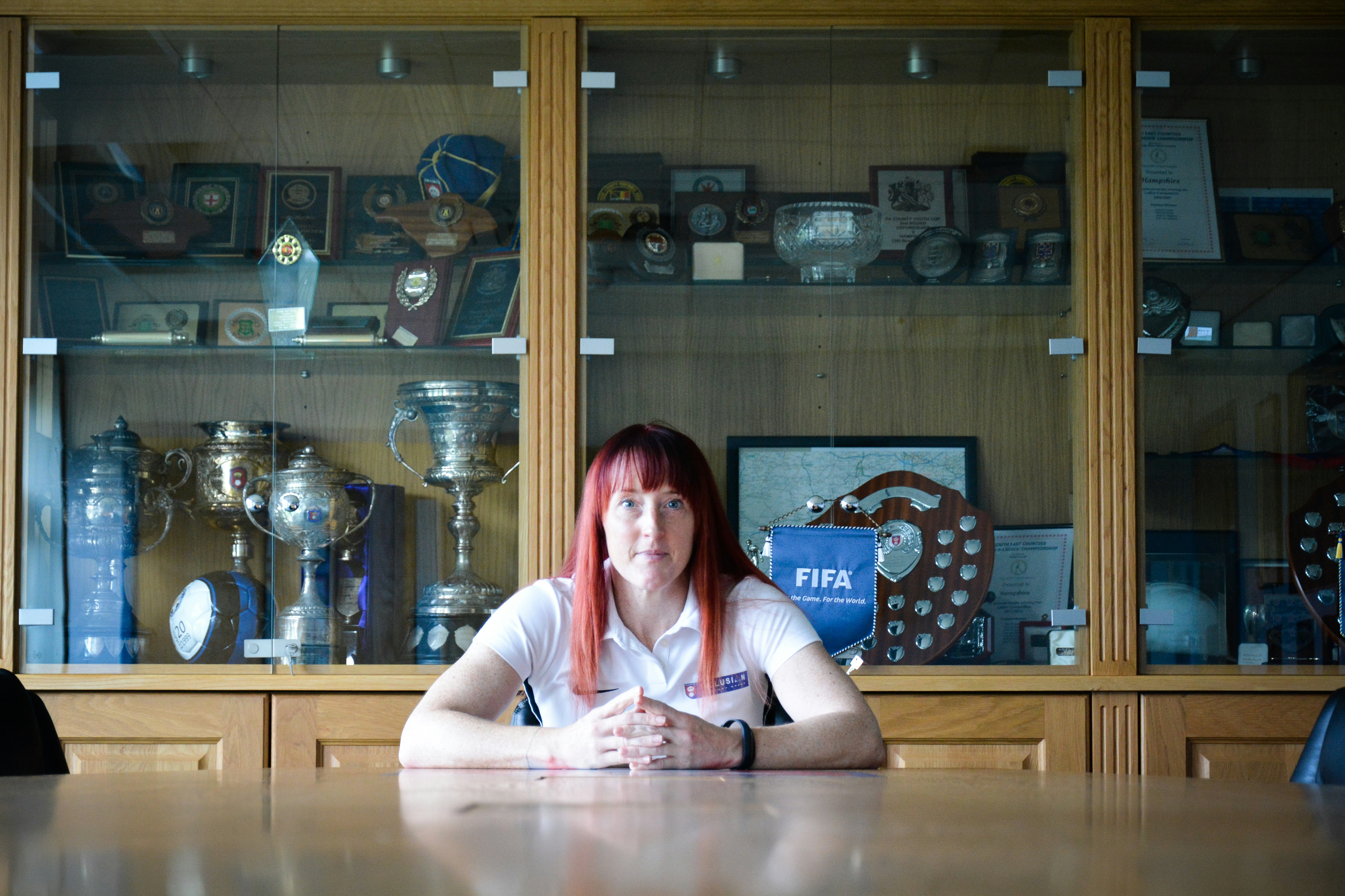 A lady sits central to the photograph gazing directly into the camera with a still face. She sits behind a wooden desk, behind is a wooden cabinet filled with trophies.