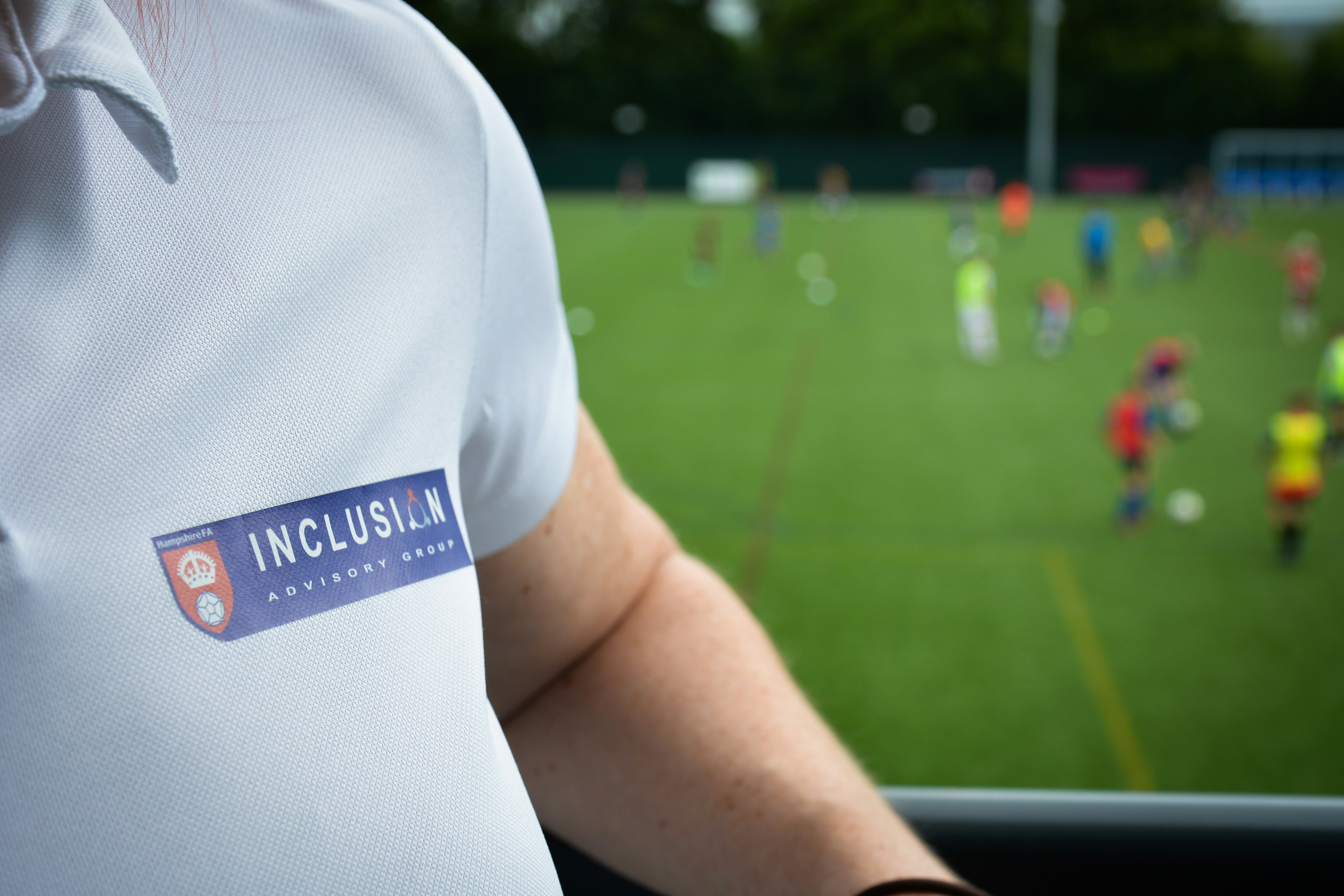 A close up of a white polo shirt with a logo which reads Inclusion Advisory Group. The logo is a blue recetange with white text. Blurred in the background shows people playing sport on a green pitch.