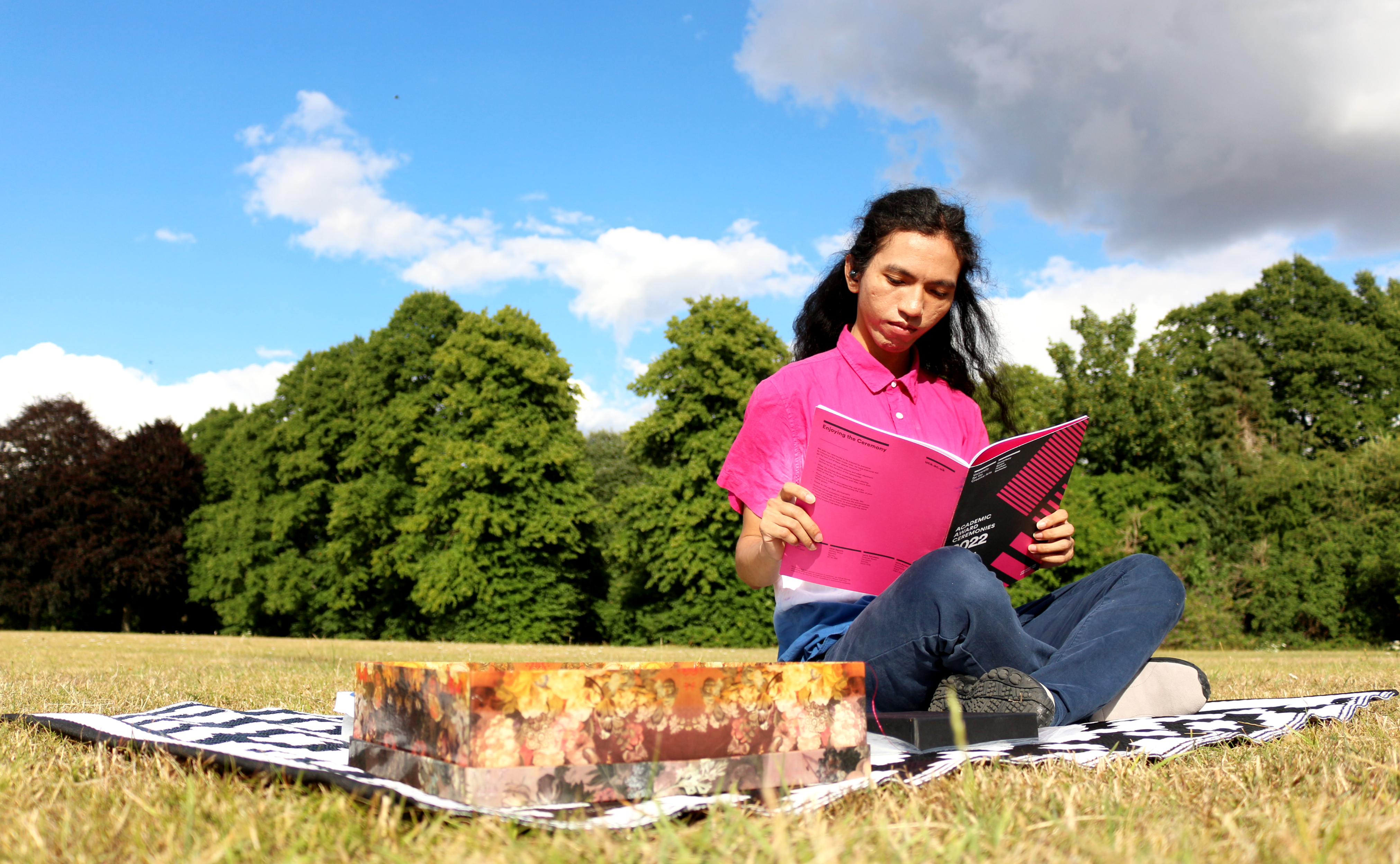 A person sits on a black and white blanket on green grass in a park. They wear a pink shirt and blue jeans. They look down reading a book.