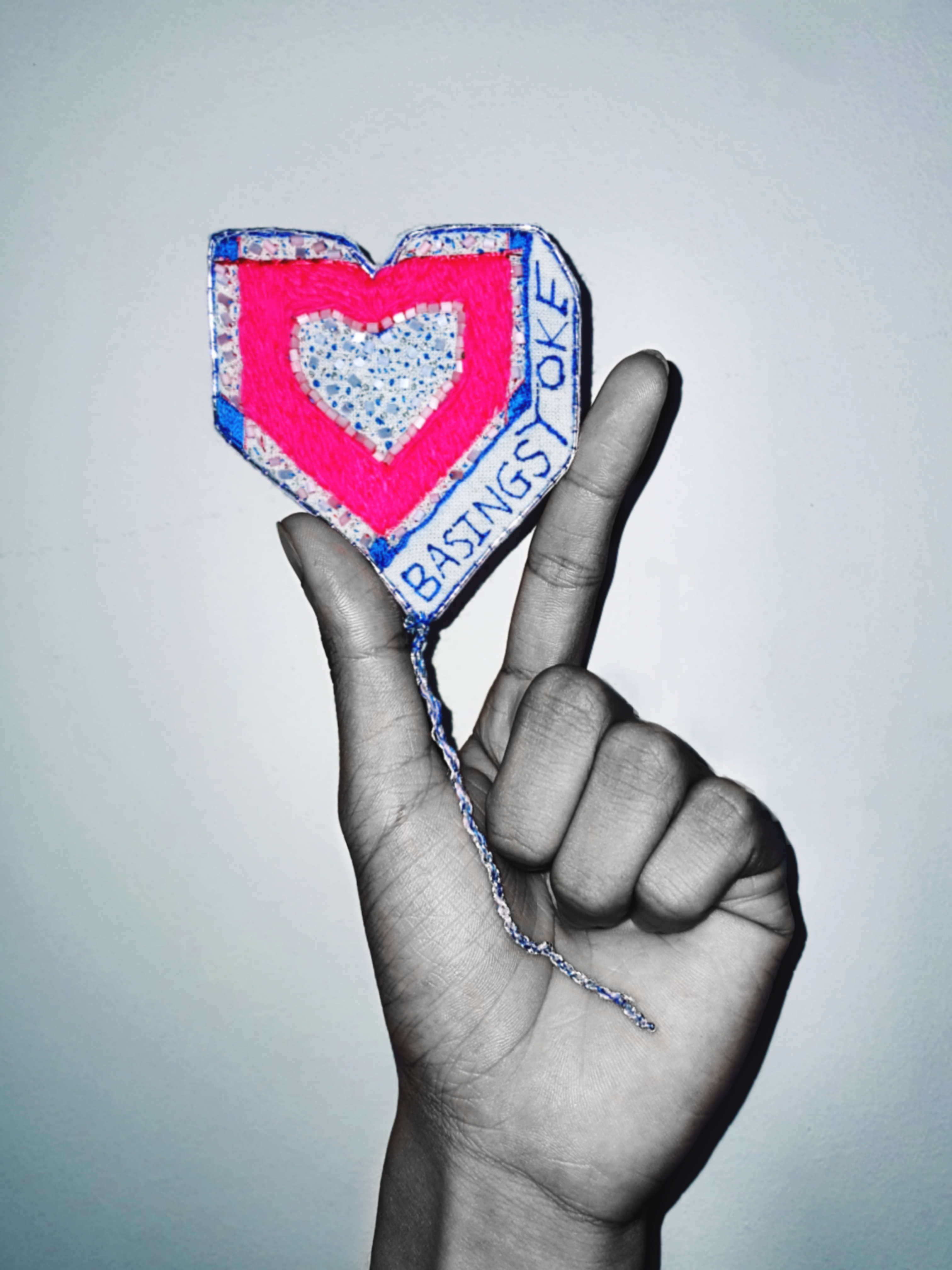 An arty image, a black and white photograph of a hand which holds a badge which is in colour. The badge reads the words Basingstoke in blue and a heart in pink thread. The badge is held between the forefinger and thumb.