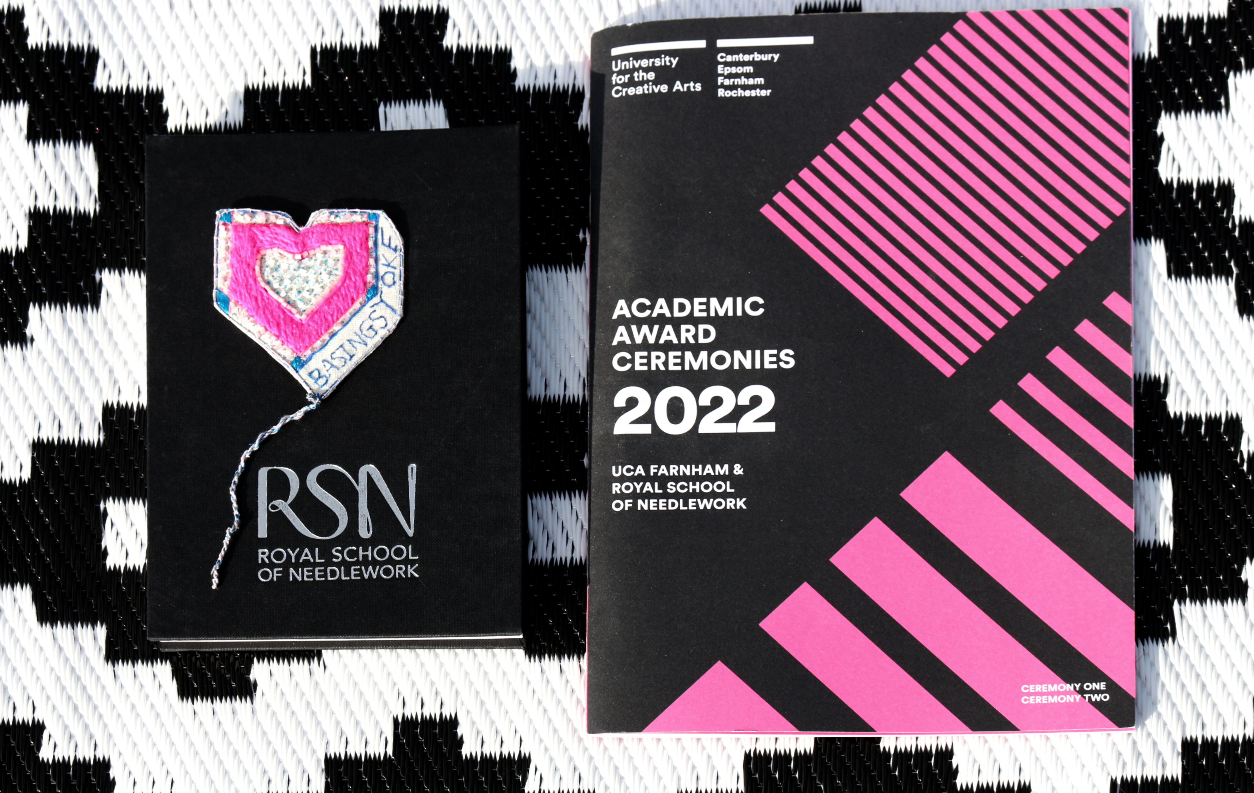 Two booklets from the Royal School of Needlework are displayed on a black and white blanket. The whole image is in black, white and pink.