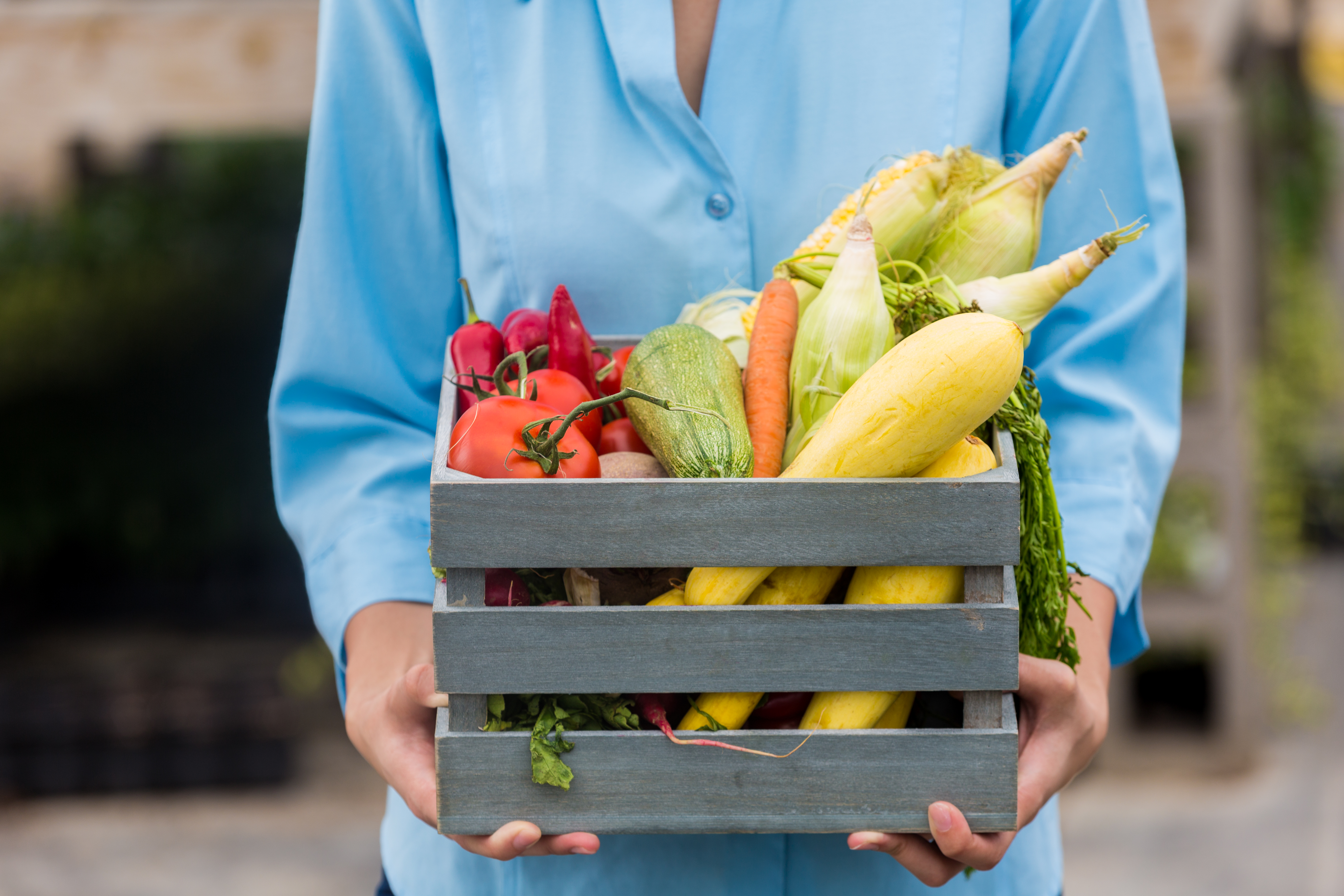 A person wearing a blue shirt carries a wooden box which contains fruit and veg.