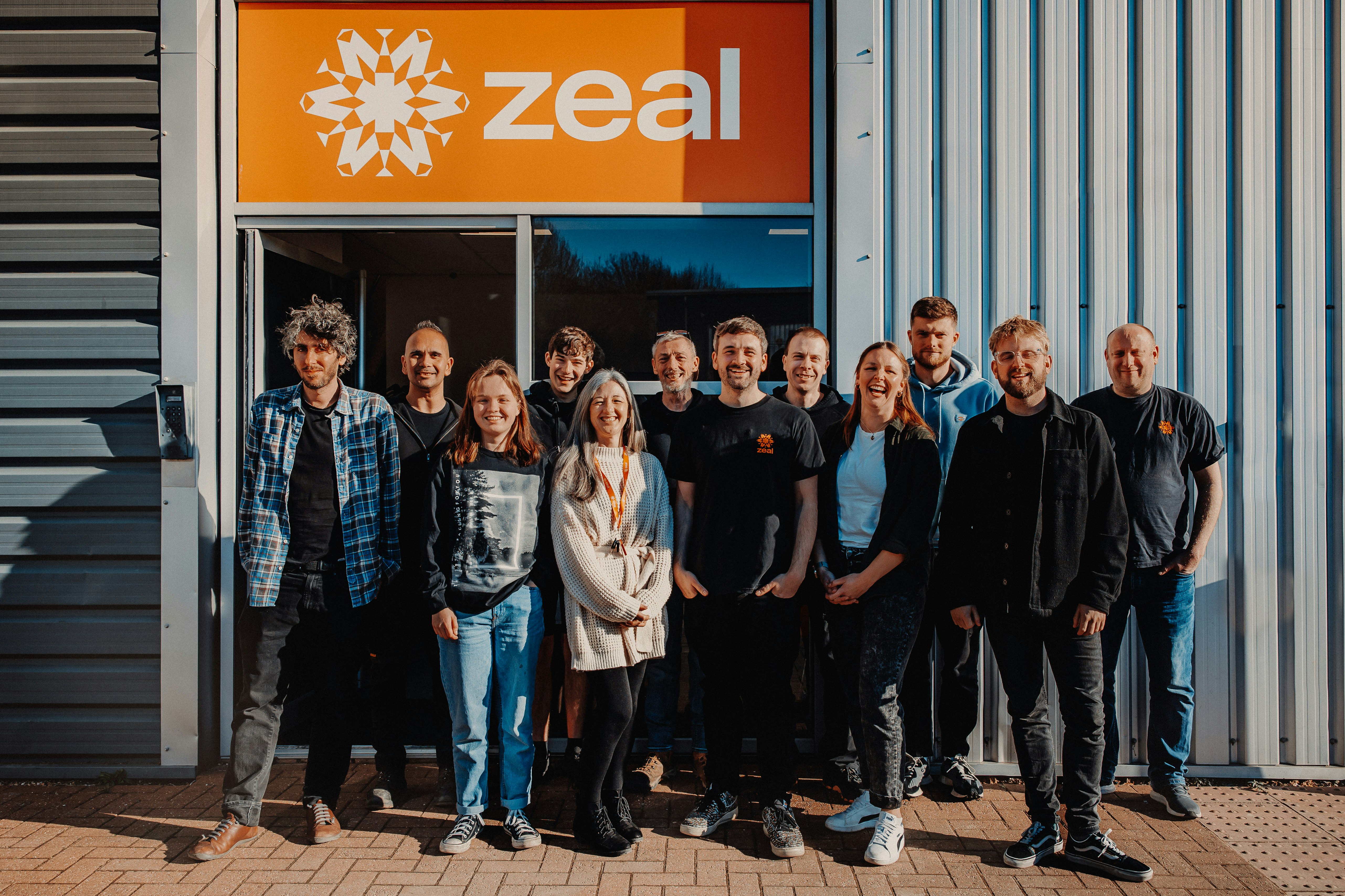 A team of 12 people stand outside the front of their office. They all smile at the camera. Behind them is a large class door, steel panels and a large orange sign with the text Zeal.