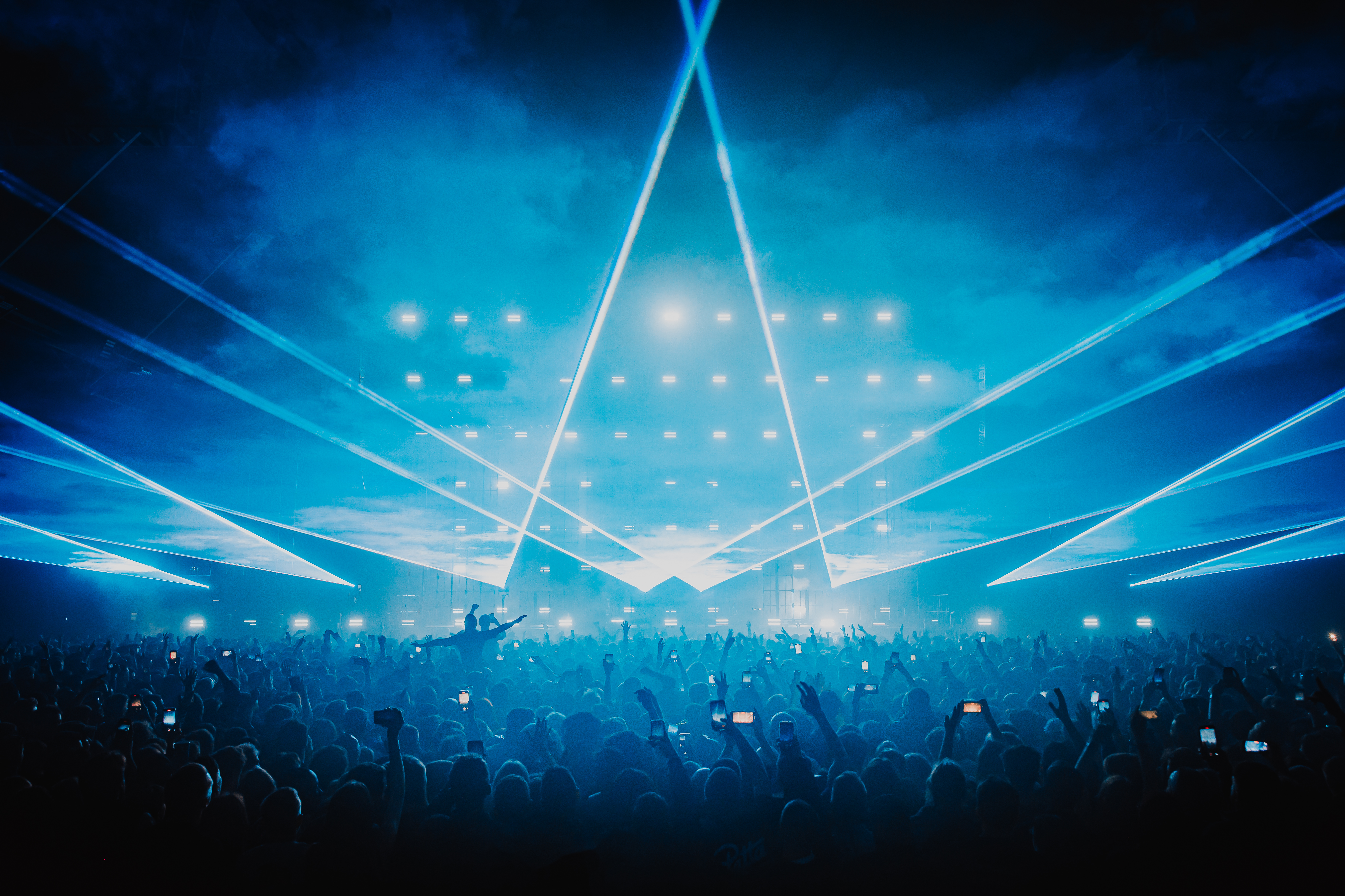 The shot is taken behind a large crowd of people with arms up looking towards a stage. The image is dark with large blue lazers lighting up the middle of the image highlight the crowds arms up in the air enjoying the performers on stage.