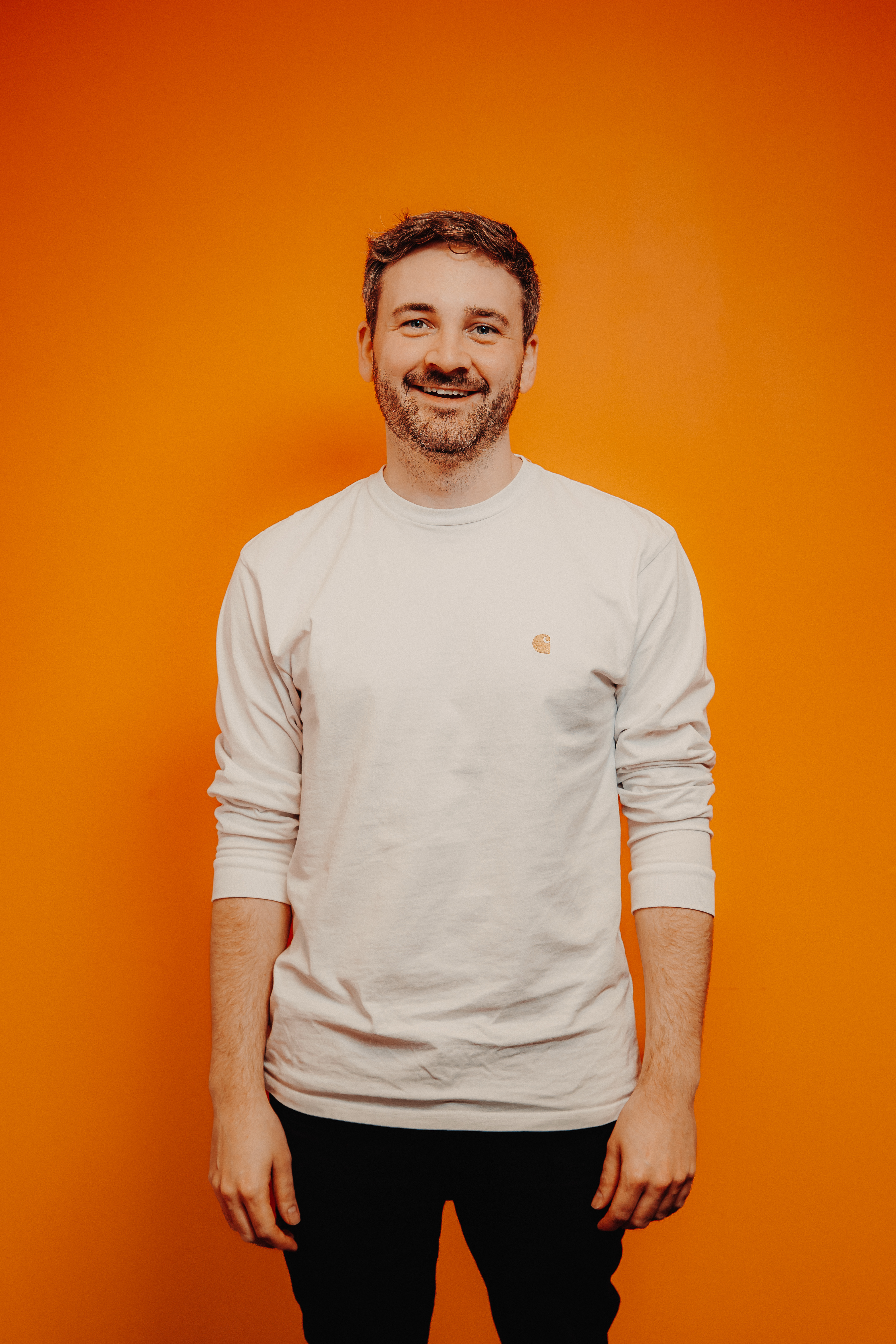 A man dressed in a cream sweatshirt and black jeans smiles at the camera, he stands infront of an orange background.