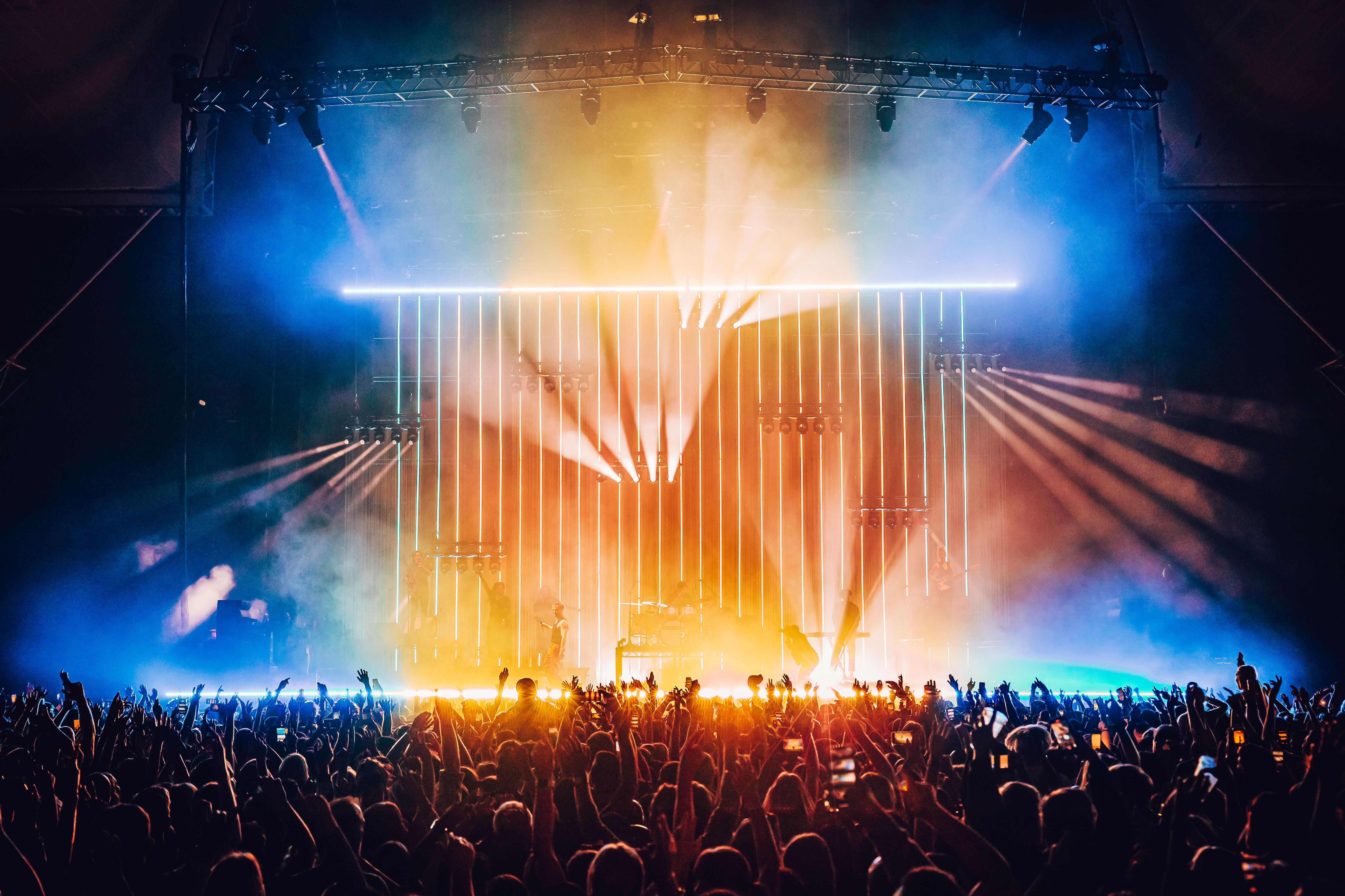 Image shot behind a crowd facing a stage. There are arms in the air, musician is on stage. The image is dark around the edges but the middle is bright with orange and blue lighting and lasers.