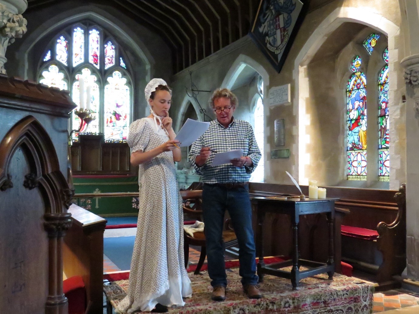 A lady stands in a dress and cap looking at a script. Next to her is a man also looking at a script. In the background you can see the inside of the church.