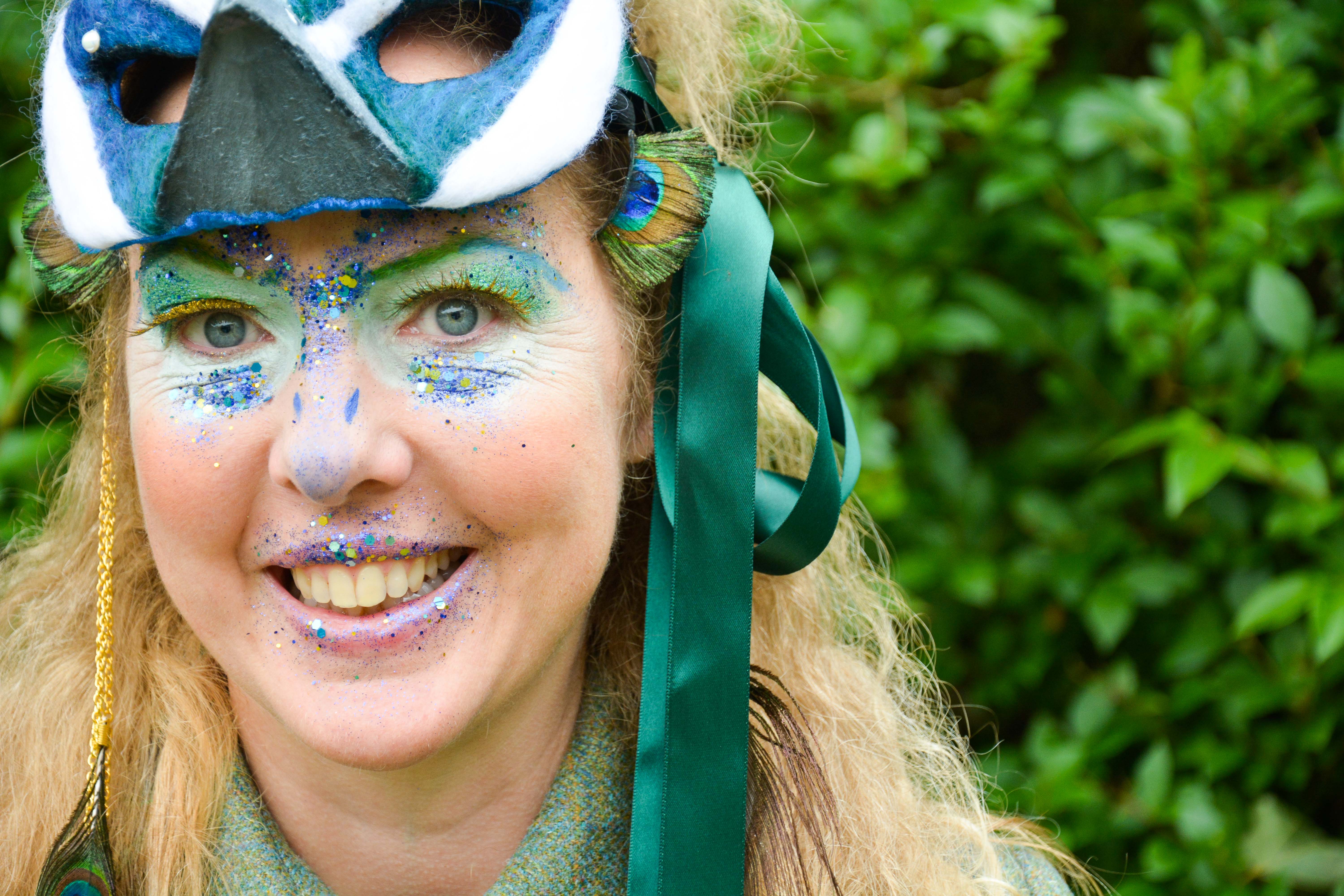 A lady wears a mask and glitter facepaint. She looks at the camera and smiles