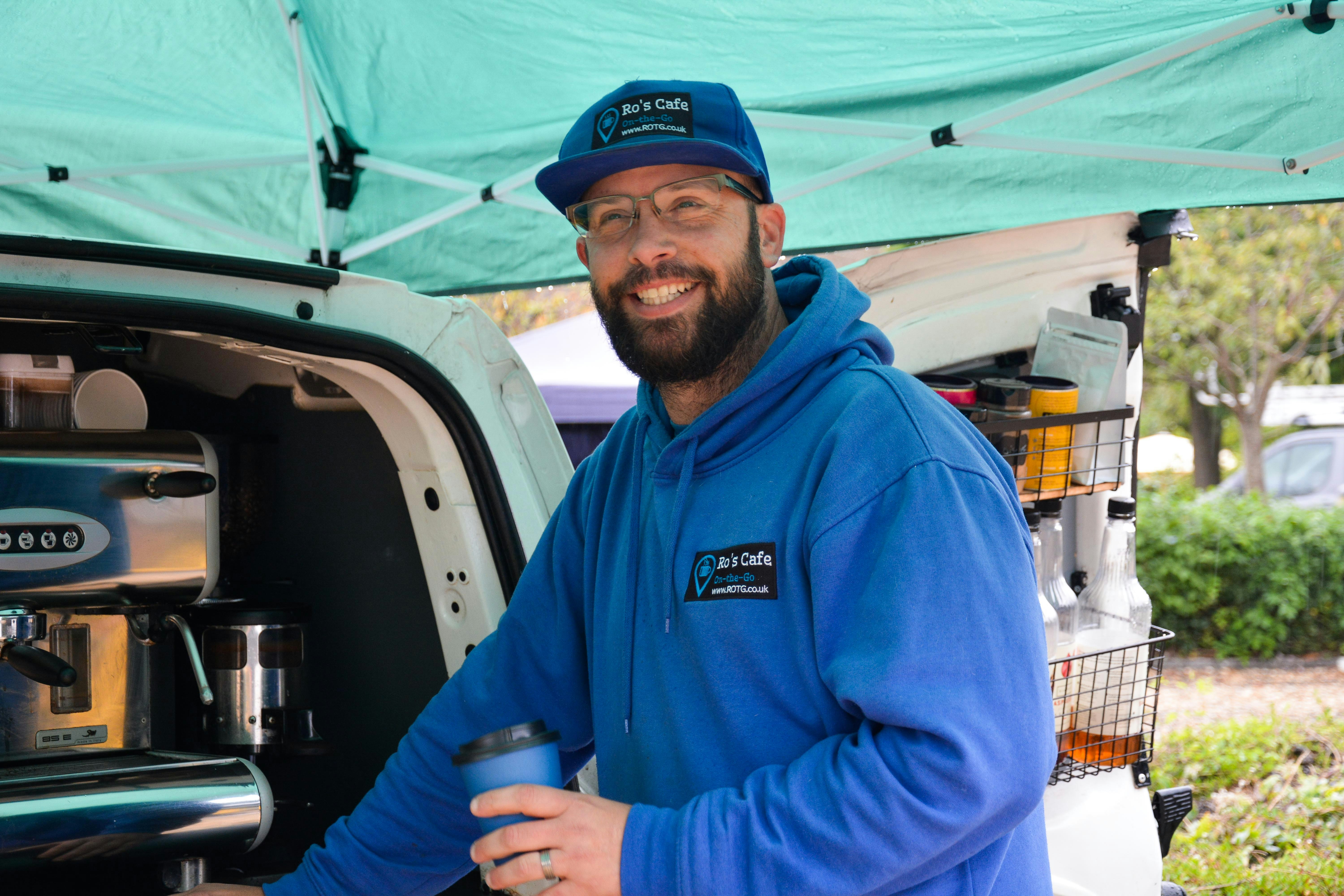 A man smiles and looks into the distance. He has a mobile coffee van behind him and he holds a coffee cup.