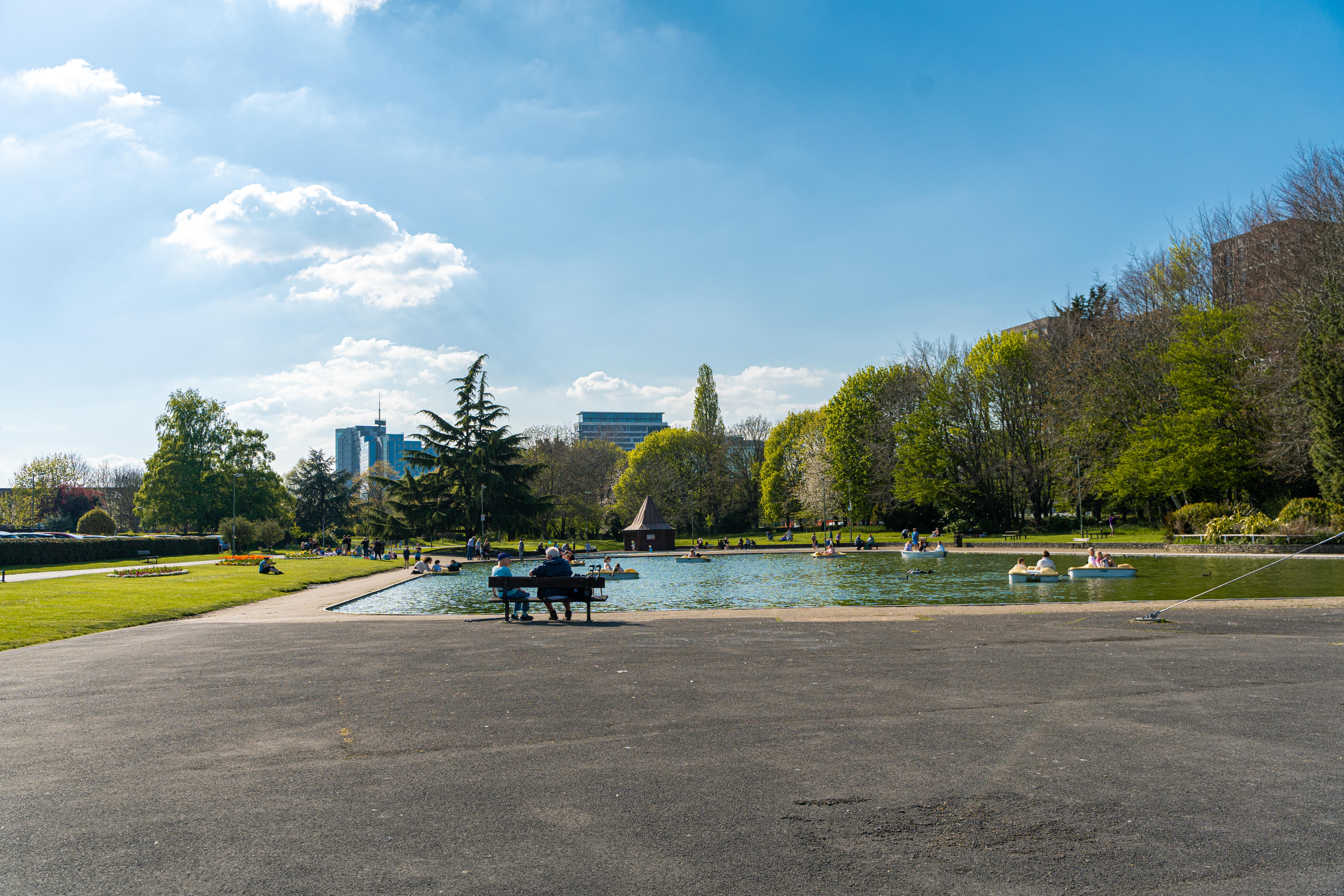 Image shows groups of people enjoying a park, boating lake and picnic areas on a sunny day.