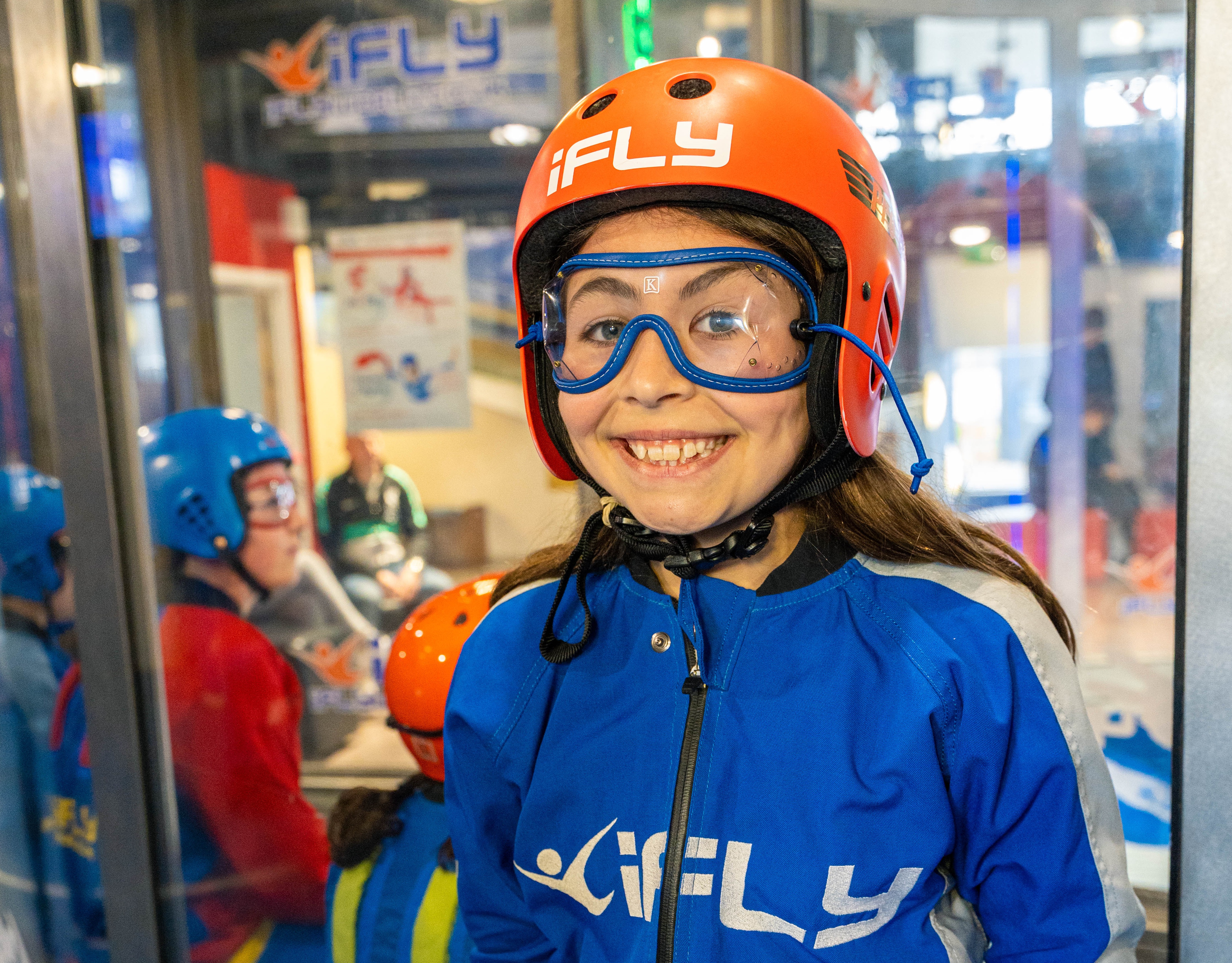 A girl smiles at the camera, she wears a light blue flight suit and orange safety helmet. She stands infront of the iFLY indoor skydiving wind tunnel.