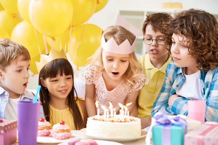 Children at a party gathered around a cake. One girl blows out the candles.