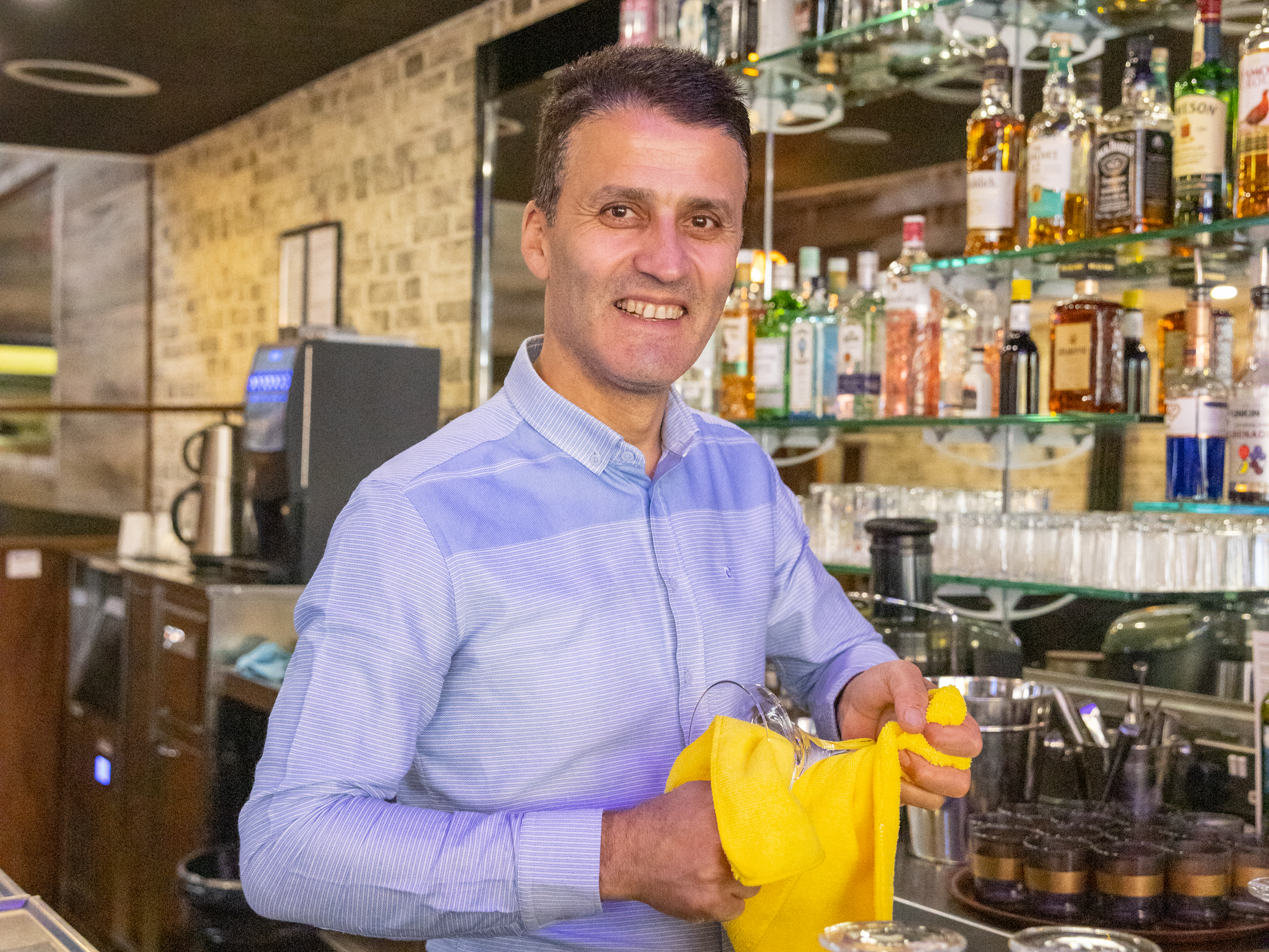 A man stands smiling and looking at the camera. He is cleaning a glass.
