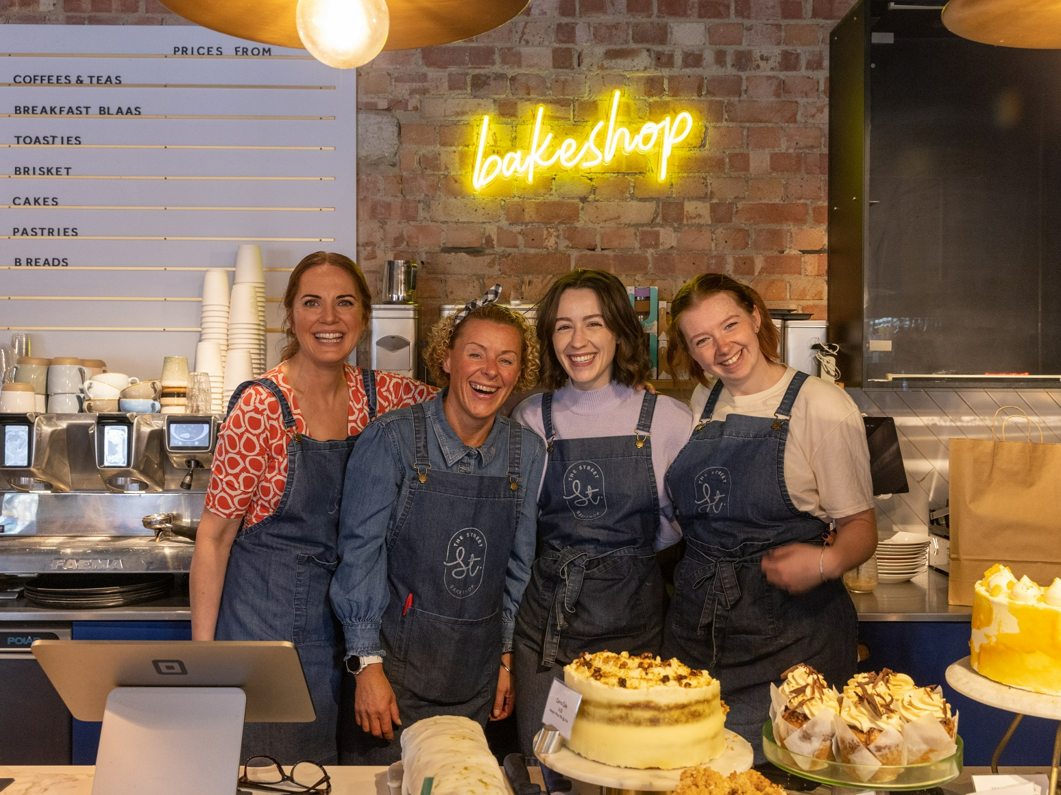 Three females wearing aprons stand together behind a counter of cakes.