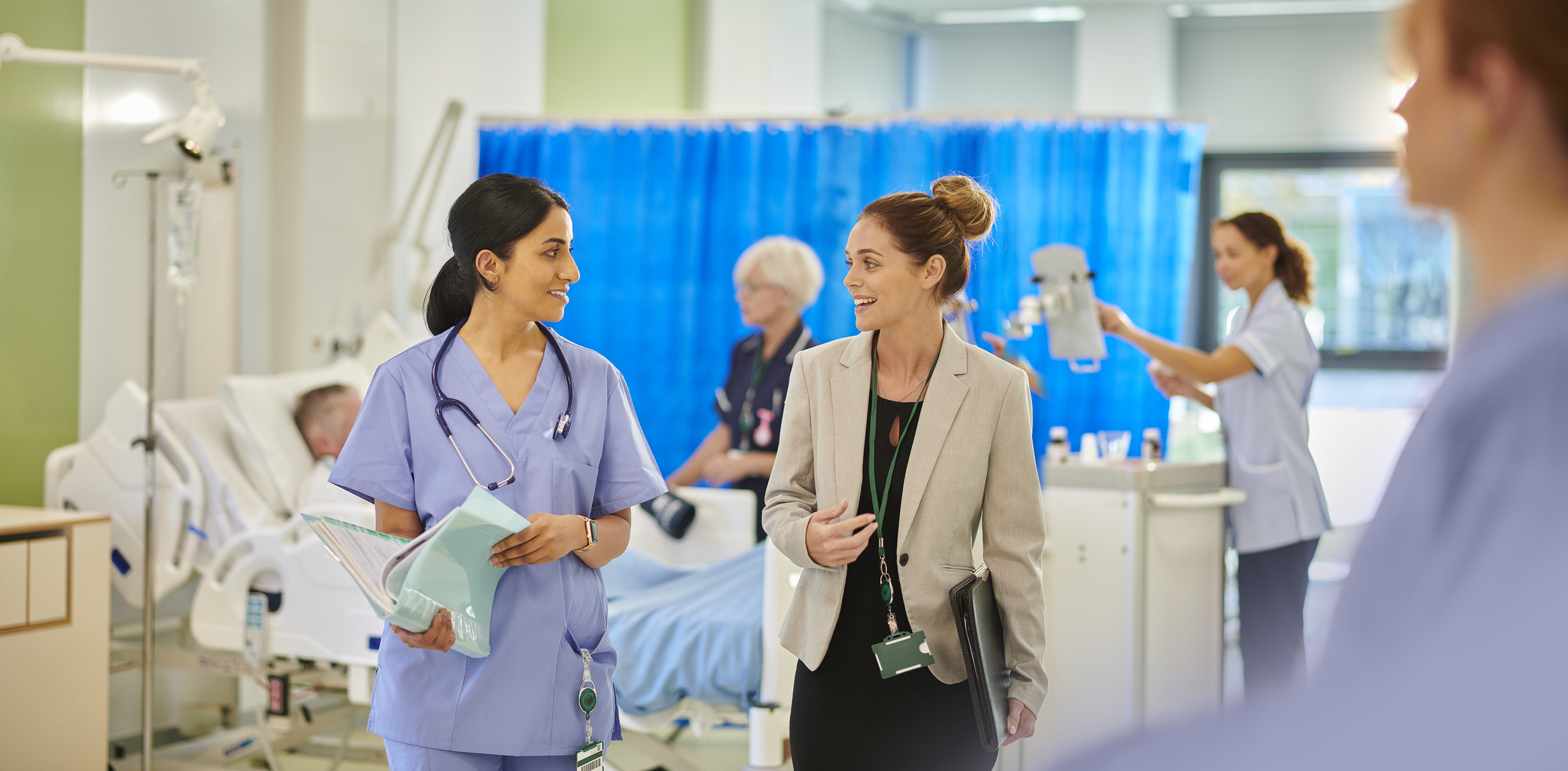 Two people talk to each other. One is dressed in scrubs with a stethascope around their neck and the other in a suit.