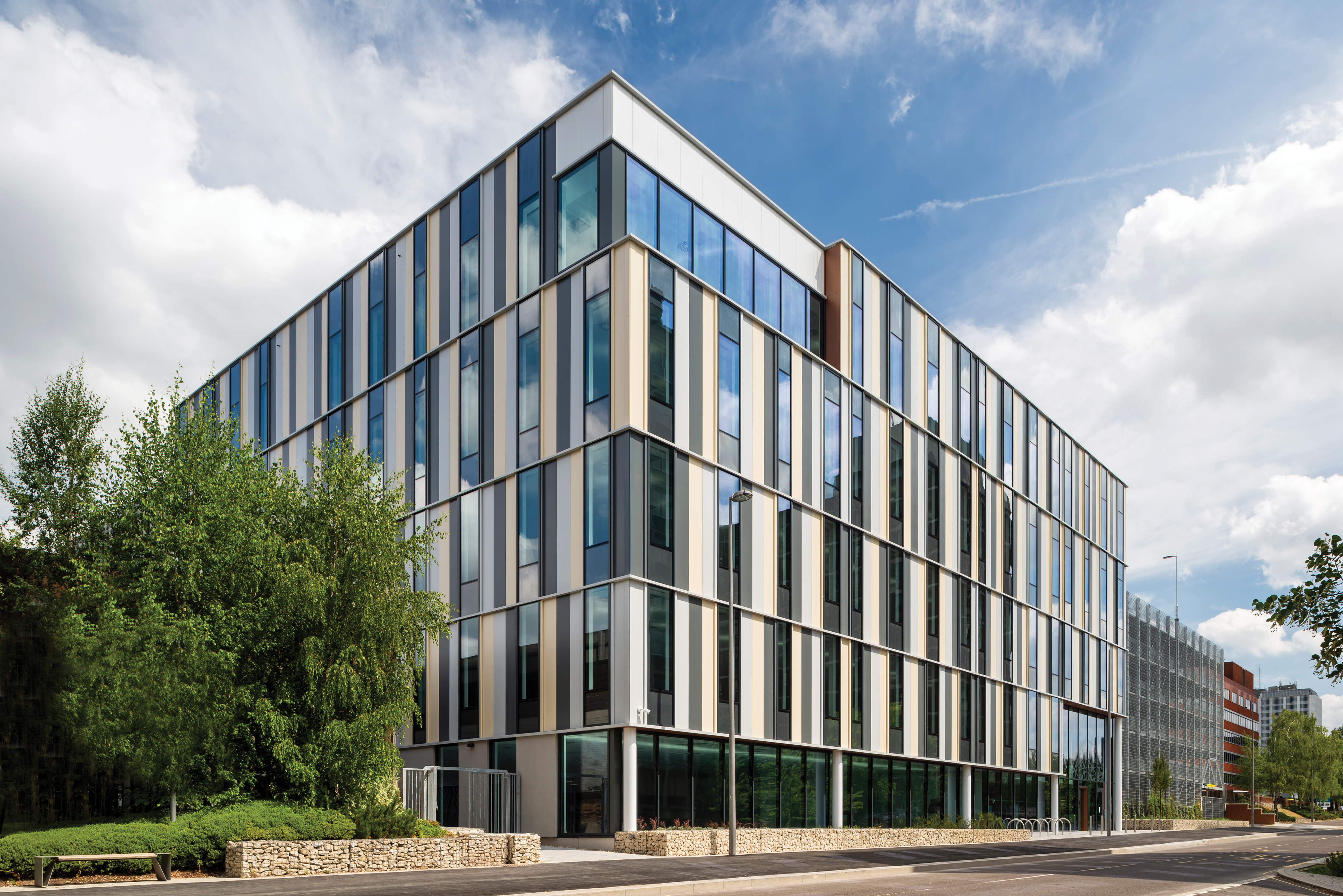A glass and panelled five storey building with a two storey glass entrance. Next to it sits a multi storey car park.