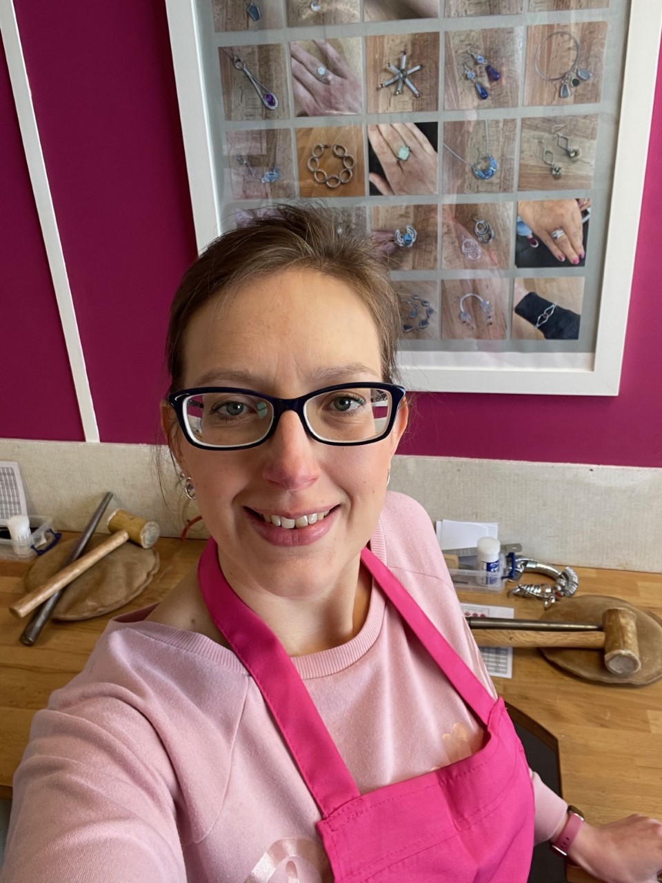 A lady looks at the camera. She wears a pink apron and sits in front of a desk.