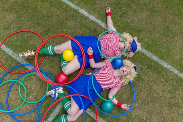 Two ladies are in sports wear and lie on the ground surrounded by balls and hoops