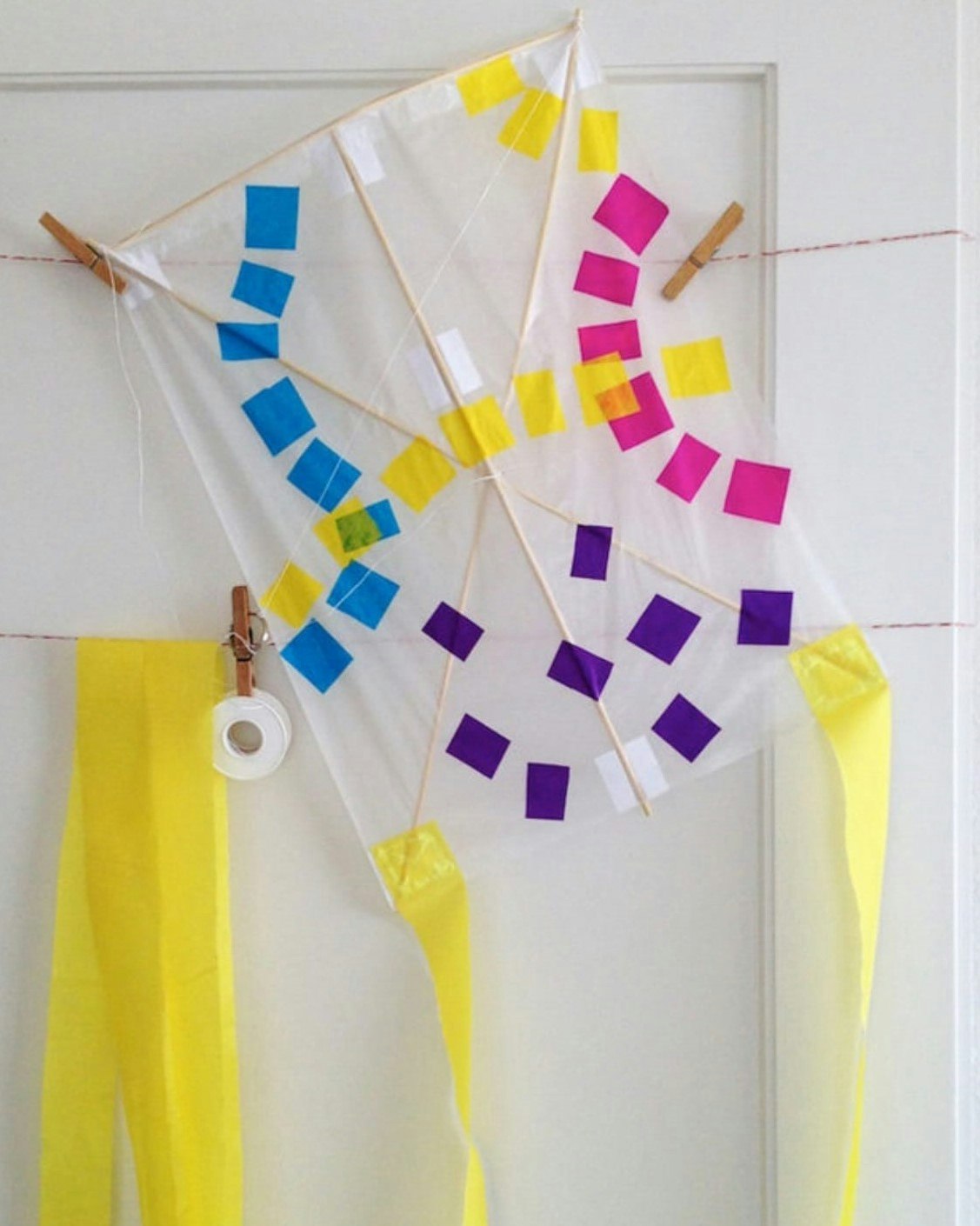 A paper kite hangs on a white doorframe. The kite is white with small blue, yellow, pink and purple blocks. Yellow ribbon hangs from the bottom of the flag.