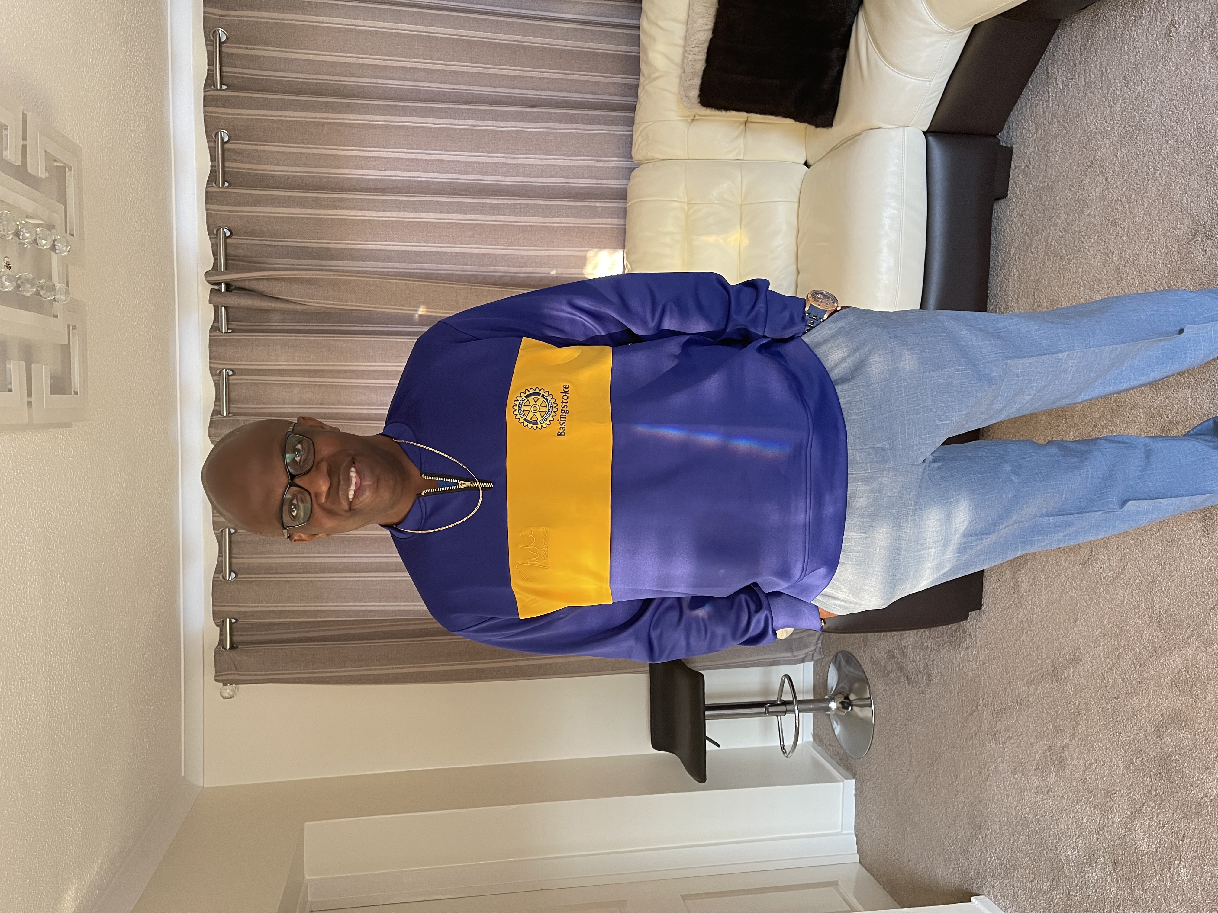 A person stands smiling at the camera, they wear black rimmed glasses, a blue jumper and demin jeans. In the background is a cream leather sofa and beige curtains which are closed.