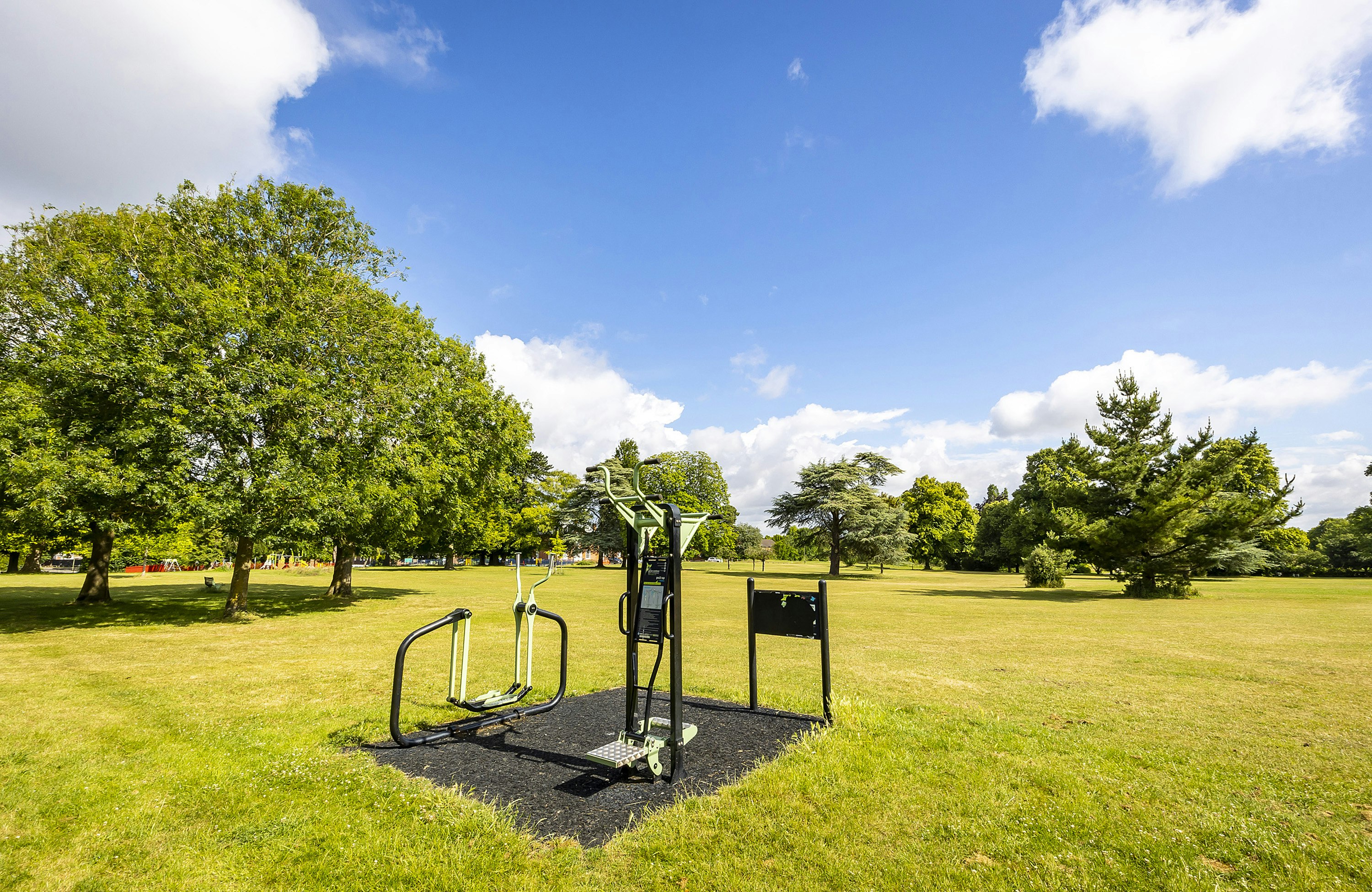 A green space within a park, gym equipment is central to the image. There are green trees in the background and bright blue sky.