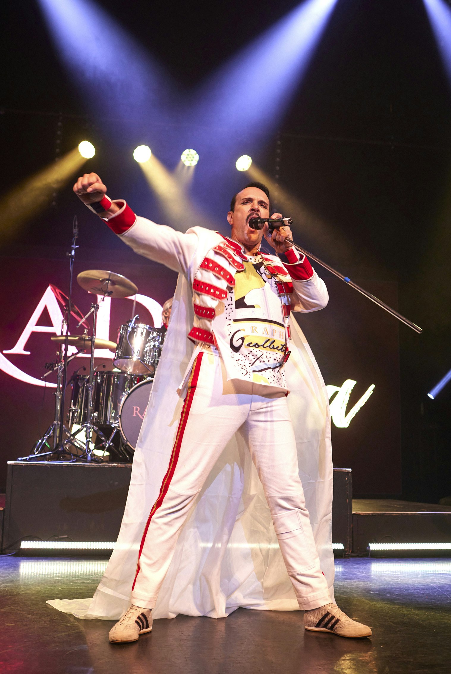 The singer of a Queen tribute band stands centre stage. Wearing a white coat with red straps and white trousers. The coat has a long white cape. The singer holds his arm in the air.