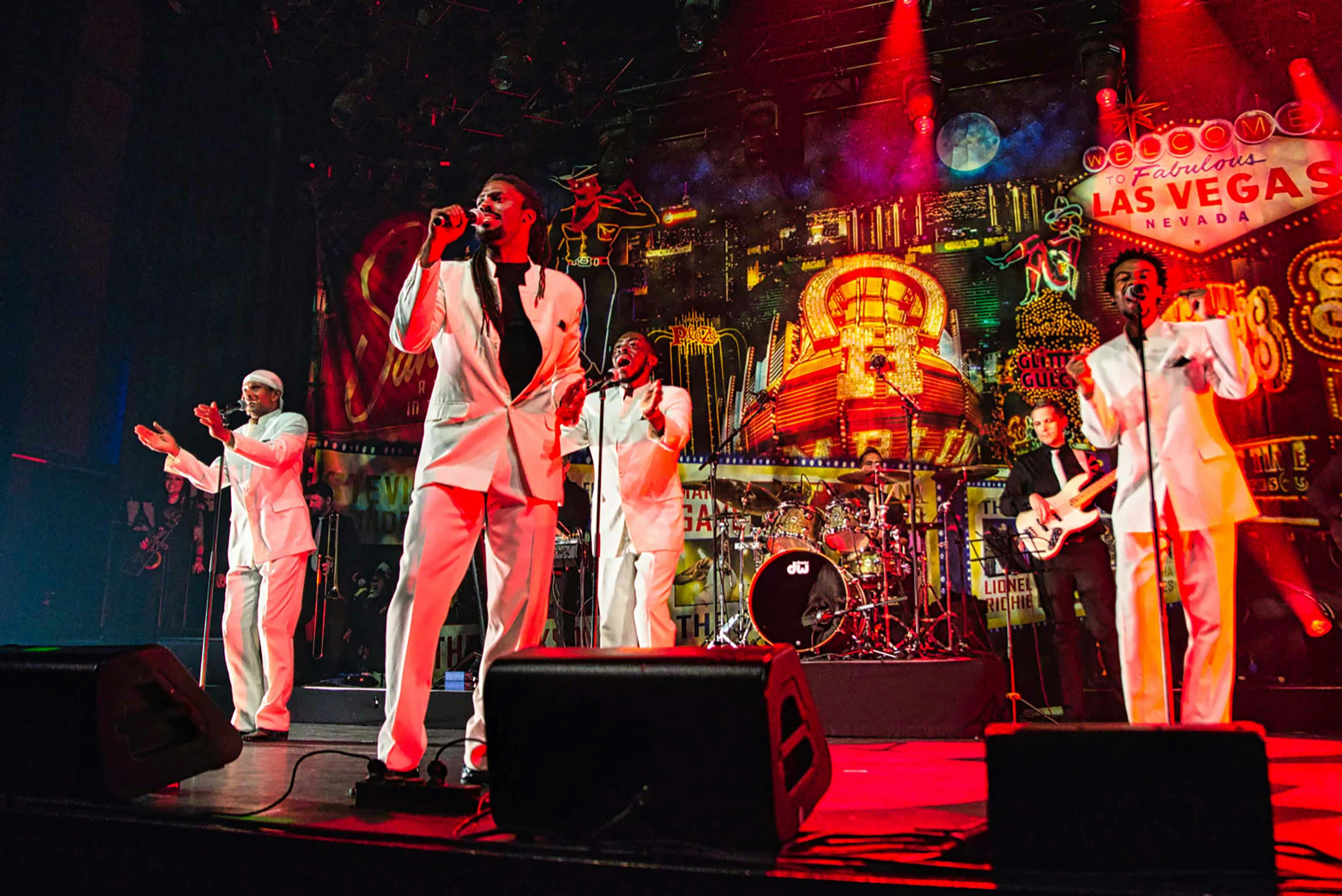 A six piece band line a stage all wearing white suits. There are four singers, a drummer and a guitarist. Las Vegas lights are the backdrop to the stage.