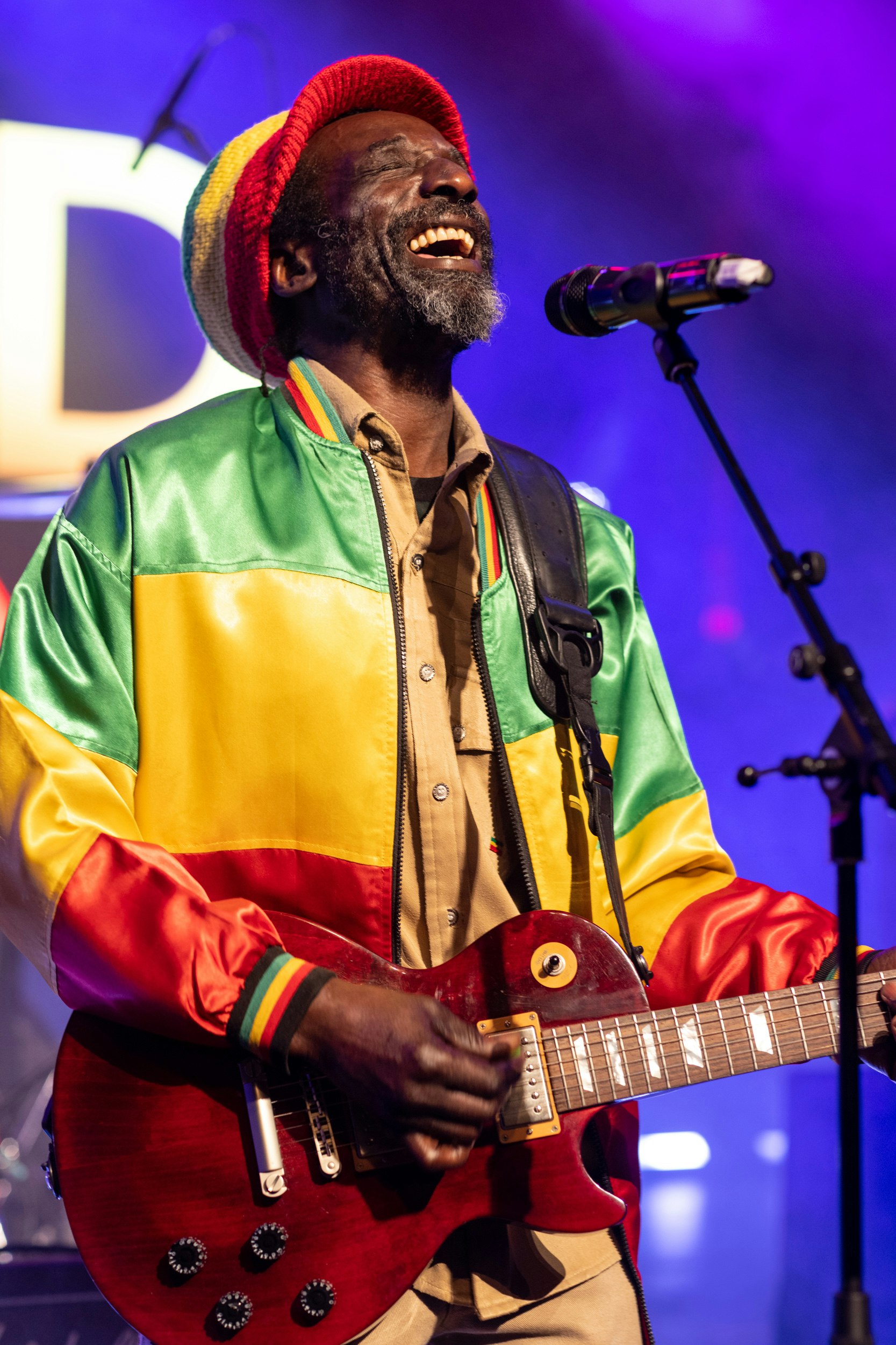 A close up shot of a Bob Marley tribute singer. He wears a red, gold and green jacket and knitted hat and holdds a red guitar. His eyes are closed, smiling while singing into the microphone.