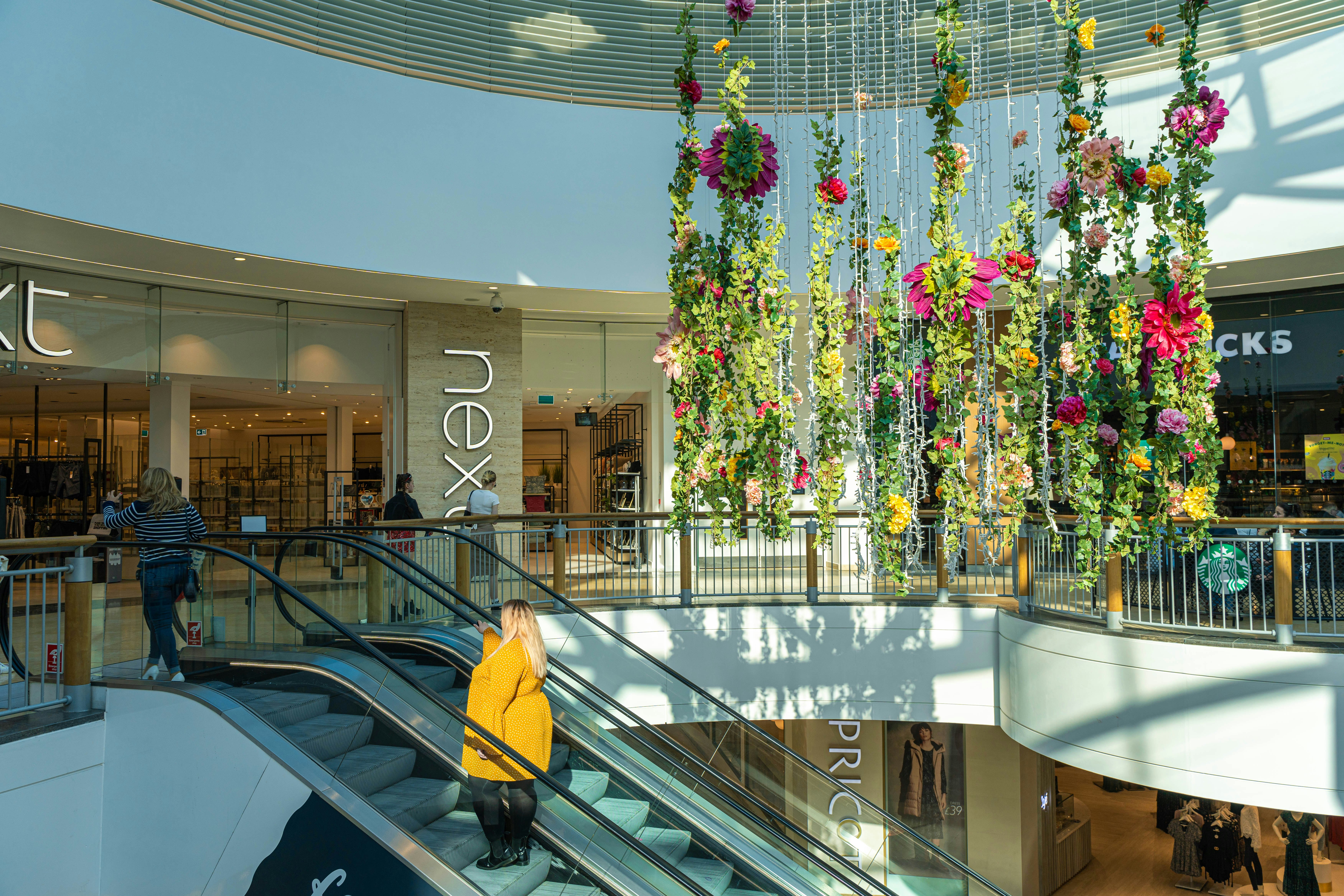 A lady wears a yellow dress and travels on an escalter inside a shopping centre. There is a large ceiling decoration with a floral arrangment.