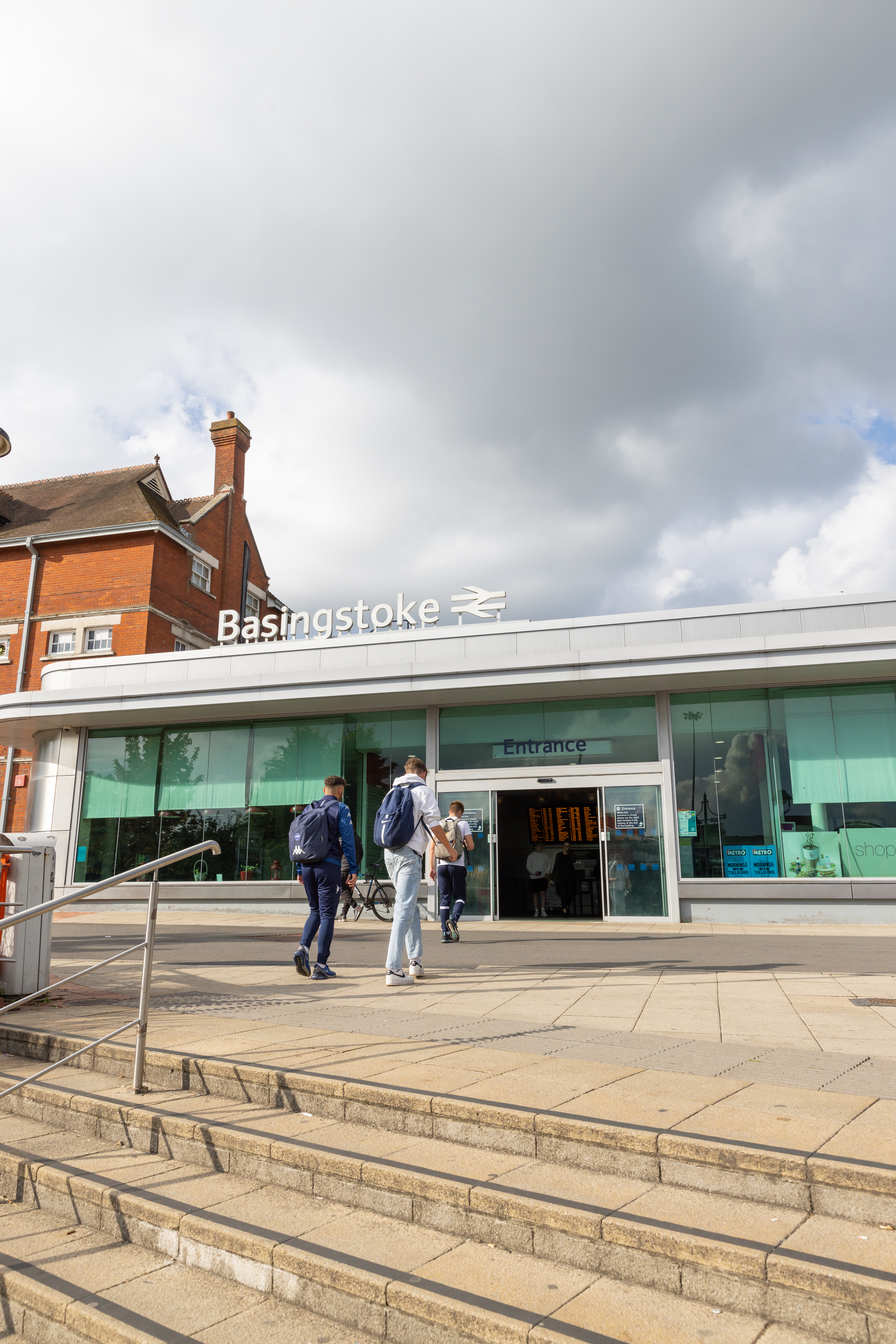 A glass fronted train station with a red brick building behind. There are two young adults walking up steps to the station.