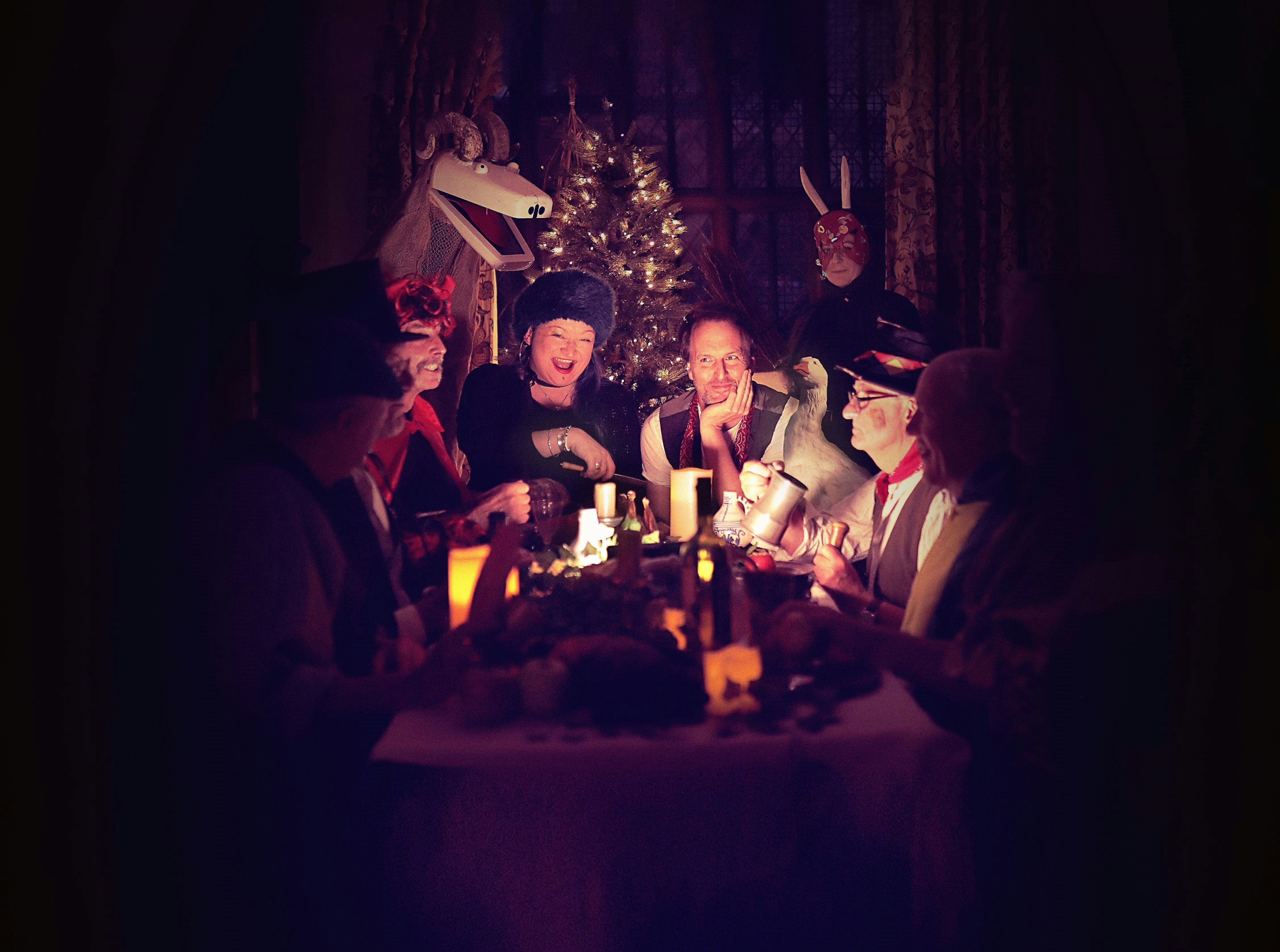 The image is dark with a table lit at the centre of the image by candlelight. A group of 6 people sit around the table laughing at eachother.